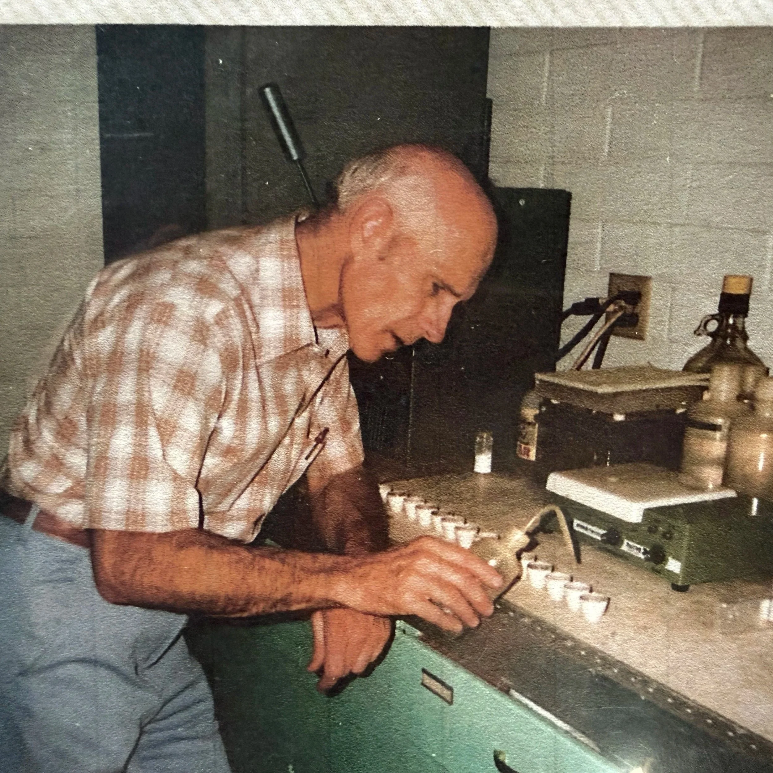 An older man with a bald head and tattoos on his arm working in a laboratory or workshop, adjusting a piece of scientific equipment on a table with various bottles and devices.