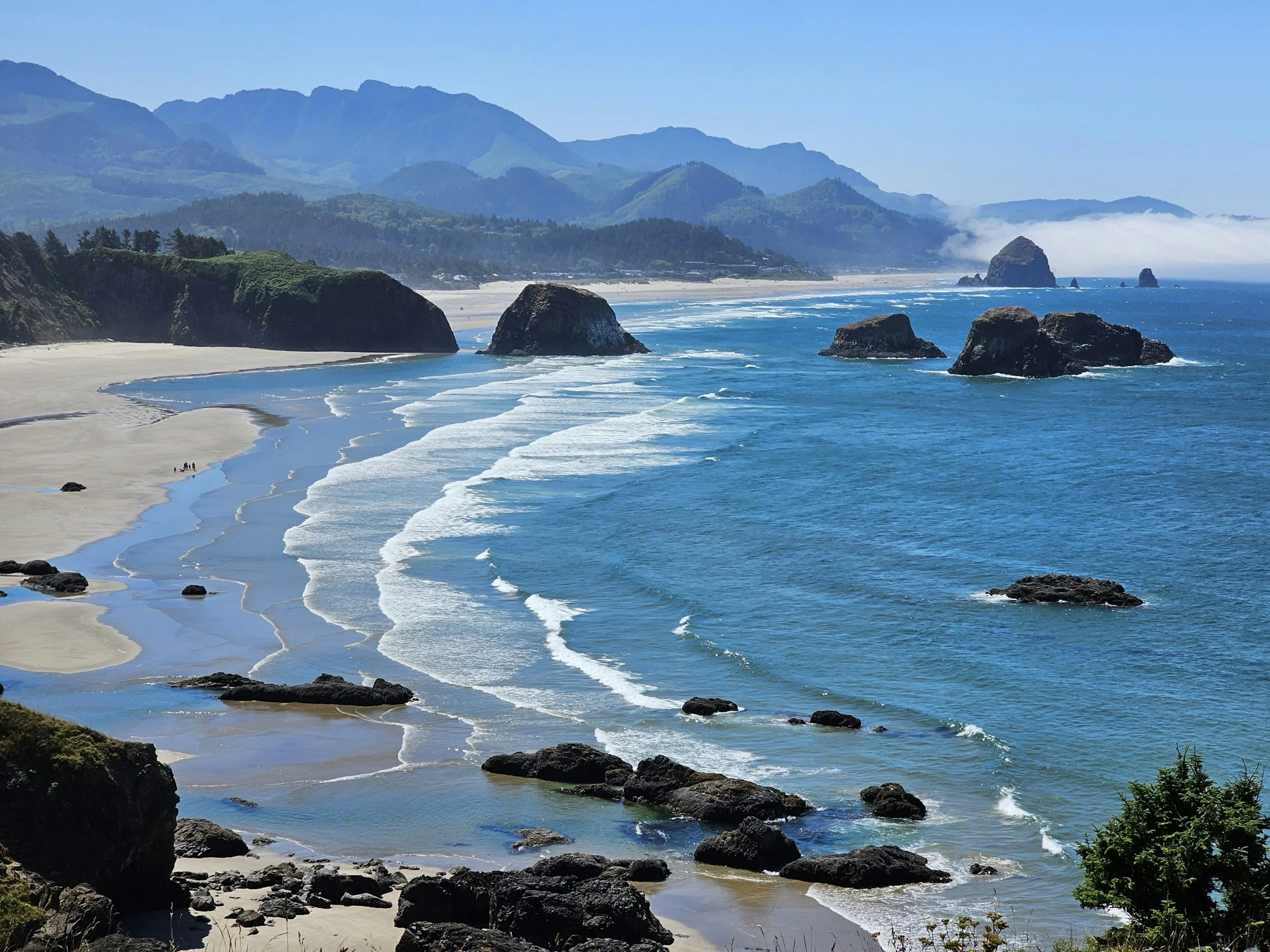 Coastal scene with sandy beach, large rocks in the ocean, lush green hills, and mountains in the background under a blue sky.