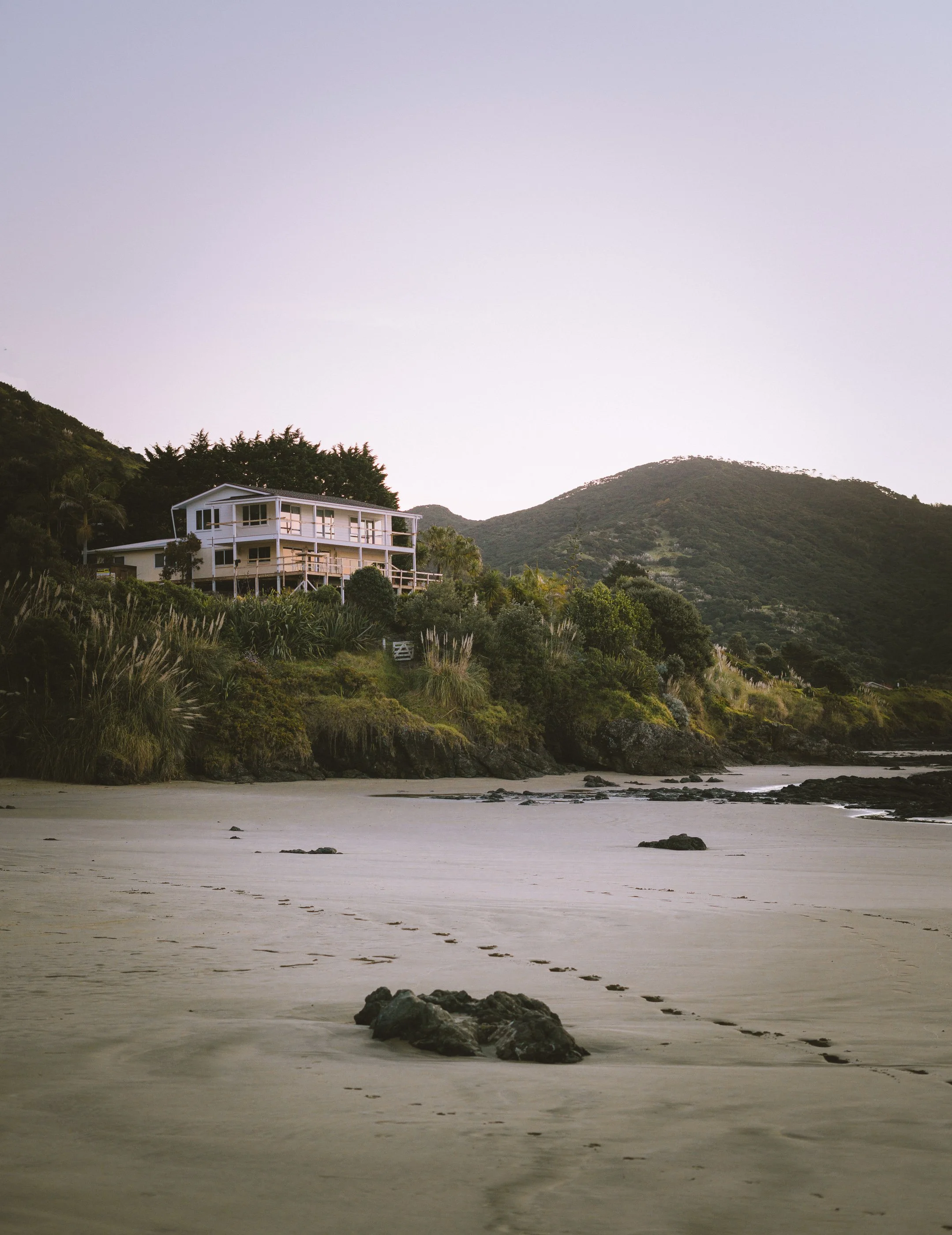 A house on a hillside overlooking a beach, with footprints in the sand and mountains in the background during sunset or sunrise.