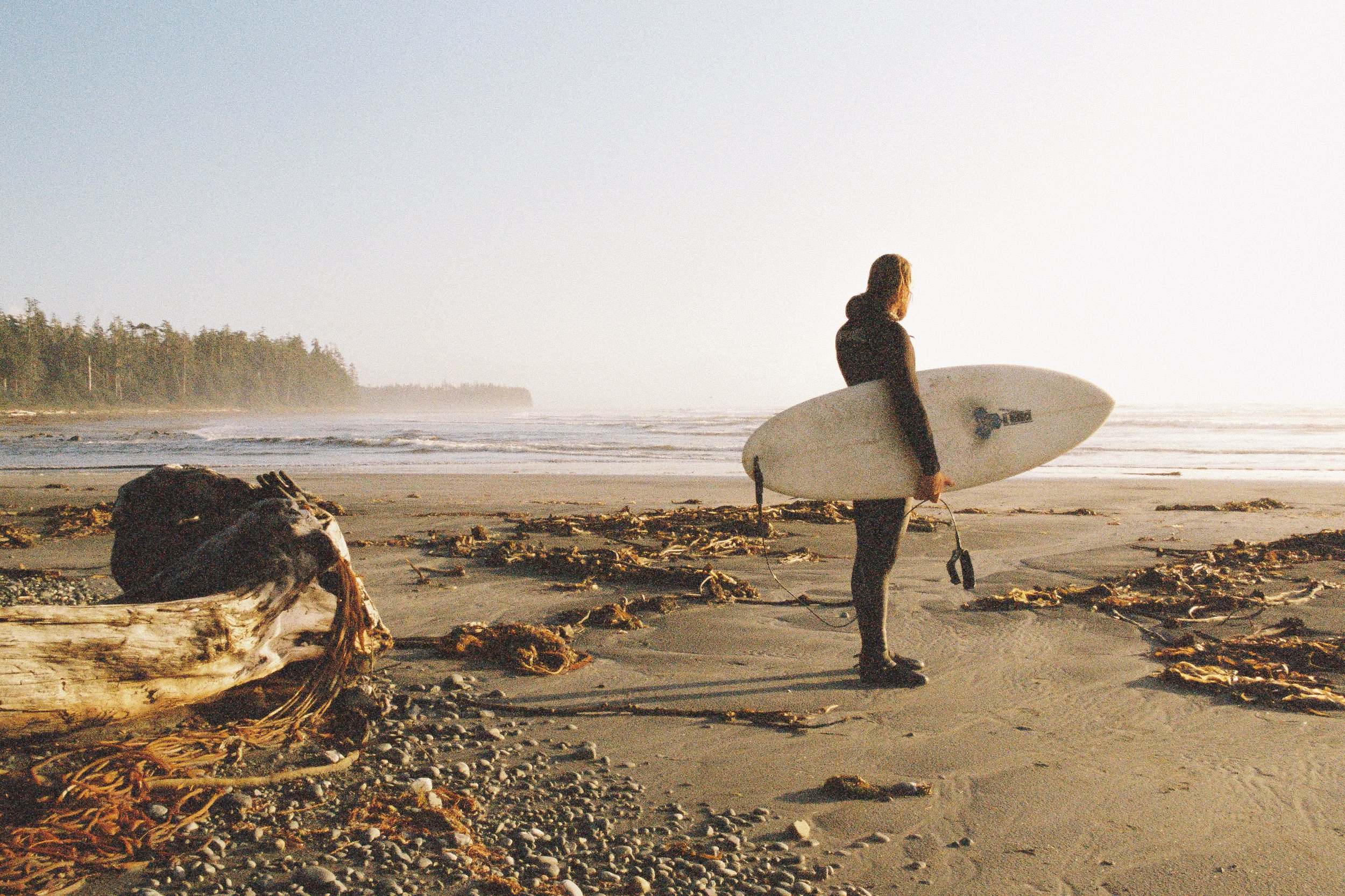 A woman holding a surfboard on a sandy beach during sunset or sunrise, with seaweed and driftwood scattered on the shore and a forest in the background.