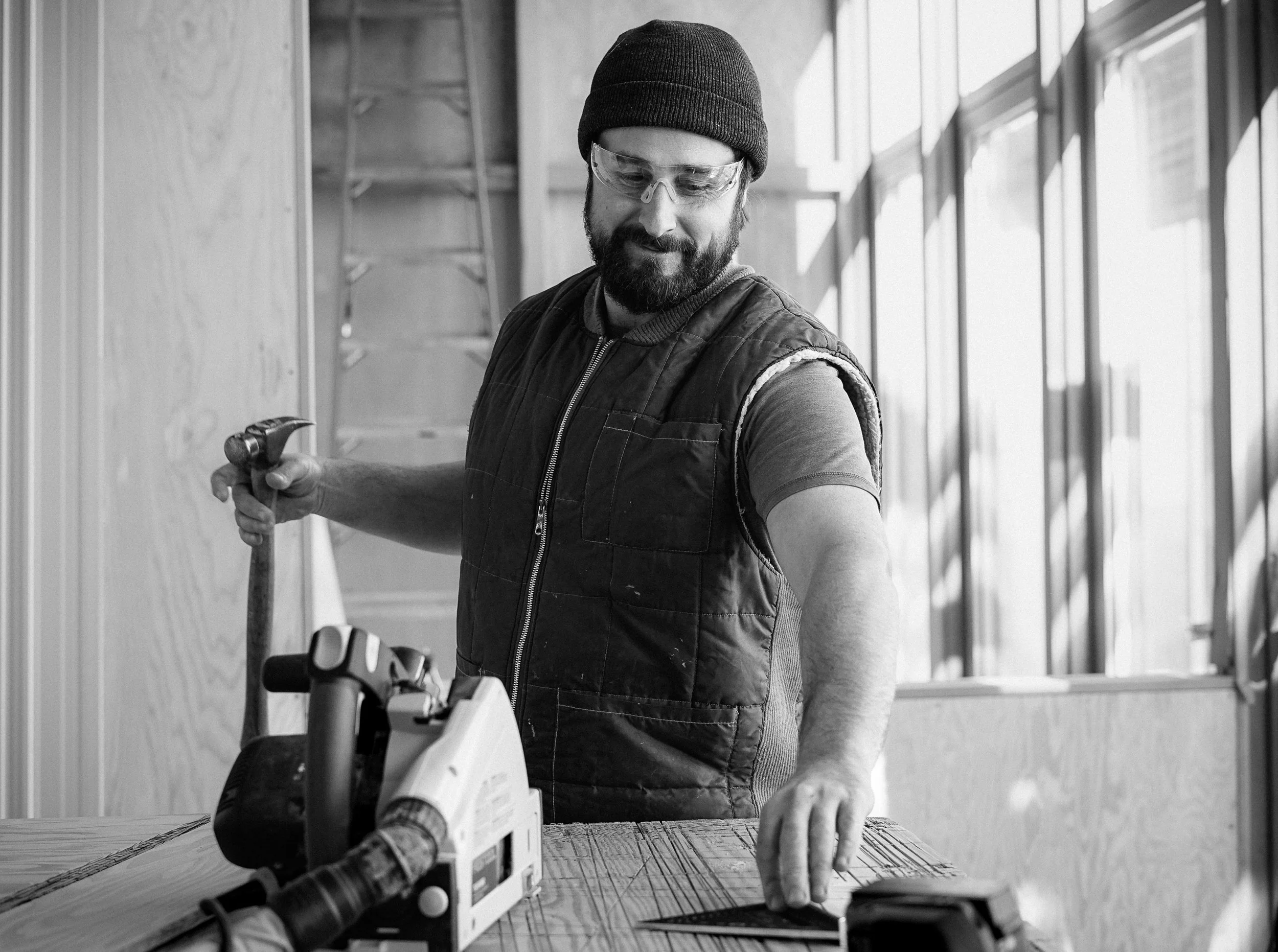 A man with a beard, wearing a beanie, safety goggles, and a sleeveless vest, is working with a hammer and a power saw on a wooden surface inside a workshop.