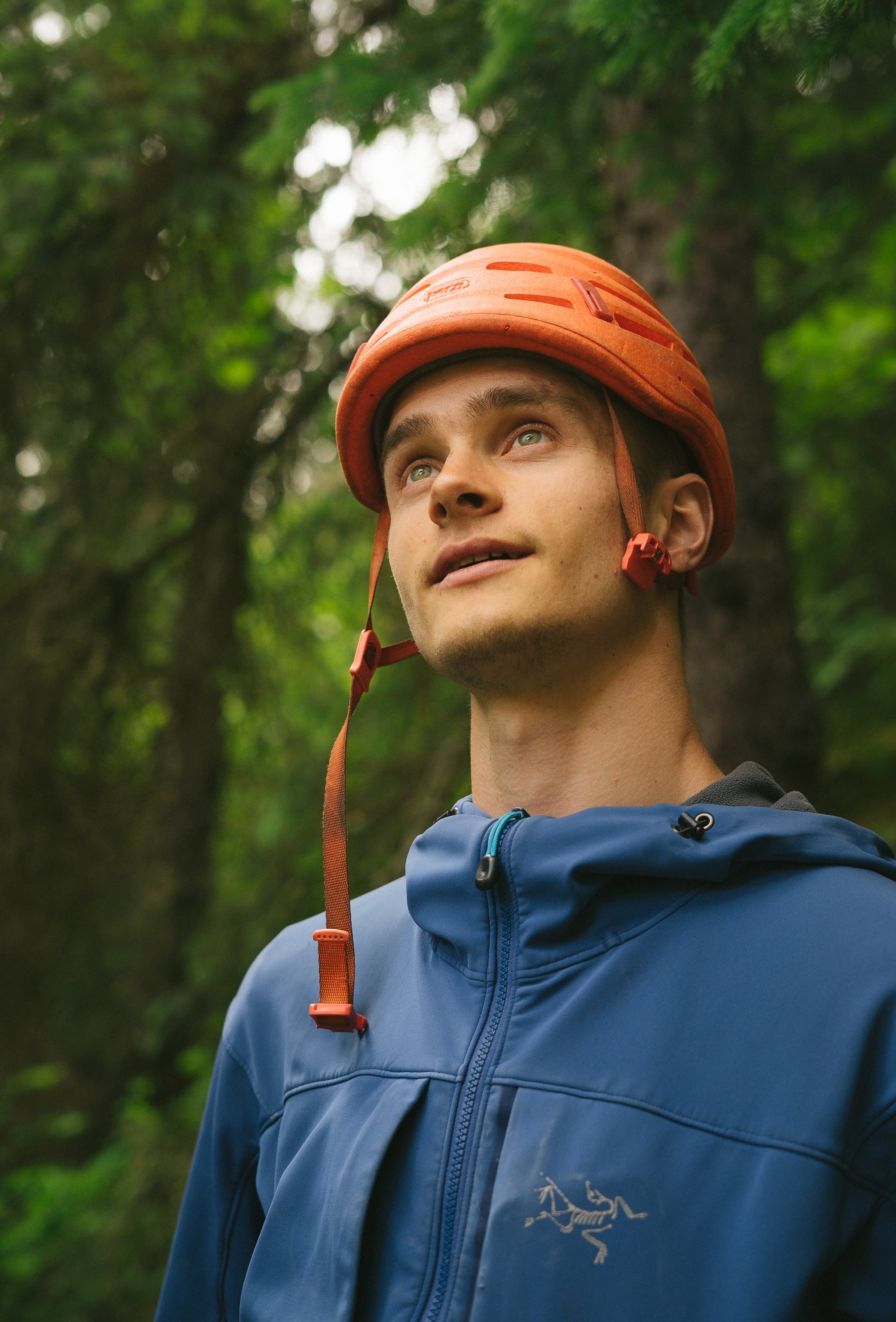A young man wearing an orange helmet and a blue jacket stands outdoors in a forest, looking upward with a thoughtful expression.