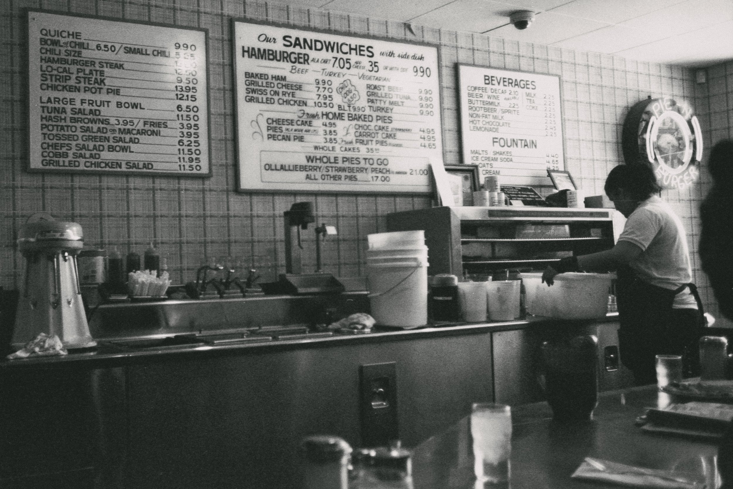 Black and white photo of a diner or cafe counter with menu boards on the wall. A person wearing a mask is working behind the counter. Various condiments and utensils are placed on the counter, and glassware is visible in the foreground.