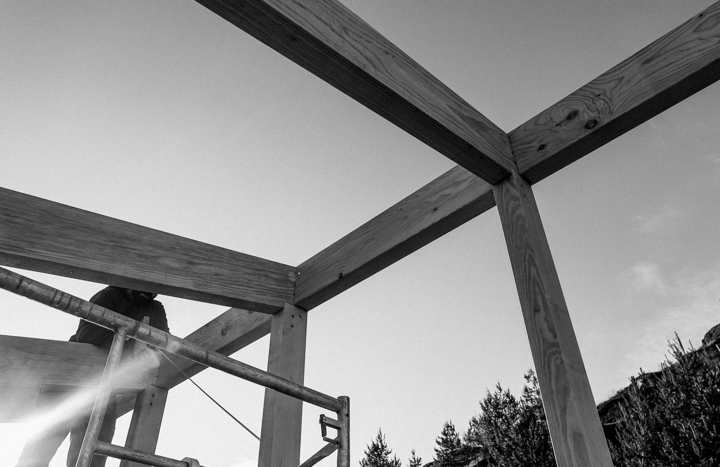 Black and white photo of a construction site with wooden beams forming a structure, a worker on scaffolding, and trees in the background.