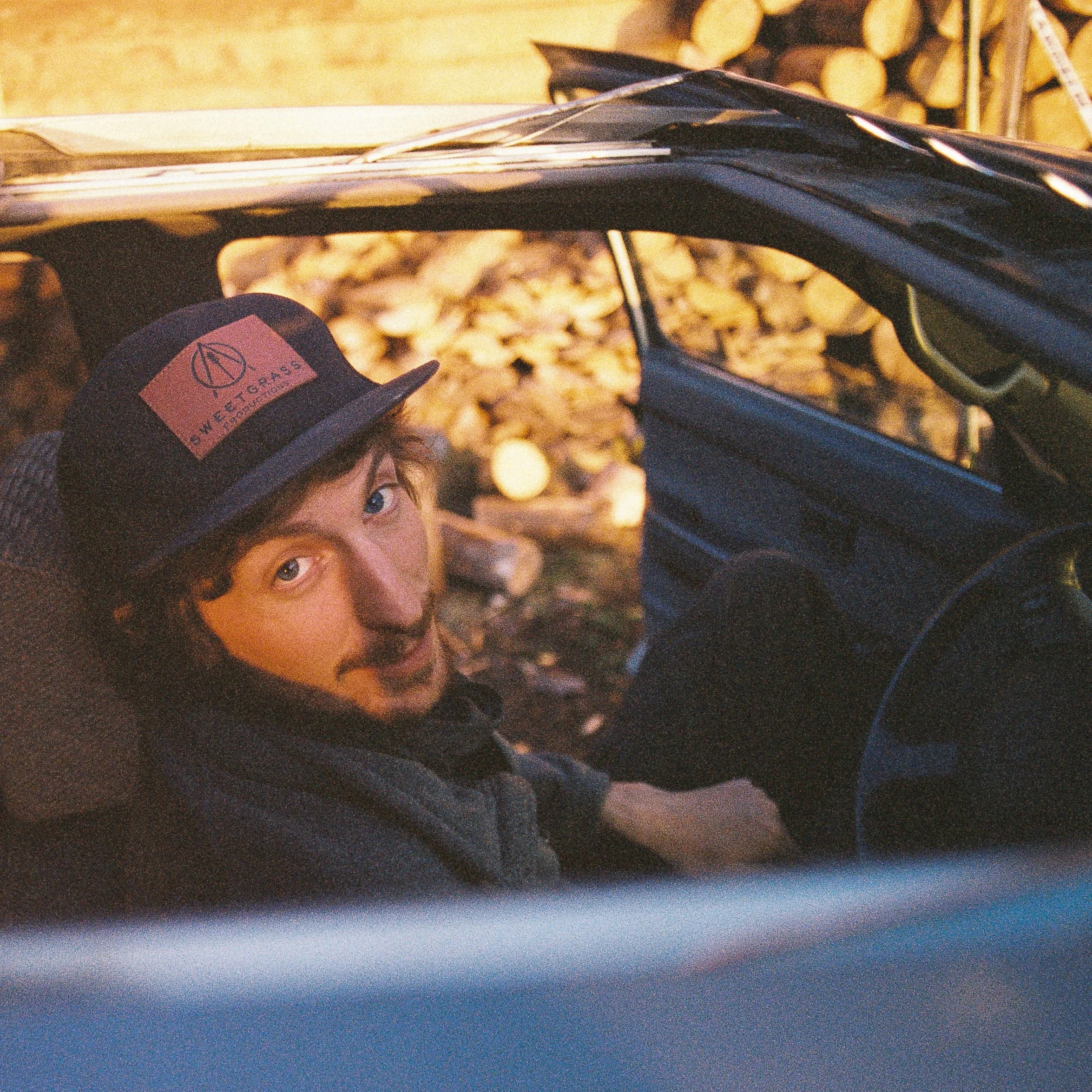 A young man with a beard and curly hair, wearing a black cap with a brown patch that reads "WETTERS" and a logo, sitting inside a vehicle with the door open, looking up at the camera. The background shows a stack of cut firewood.