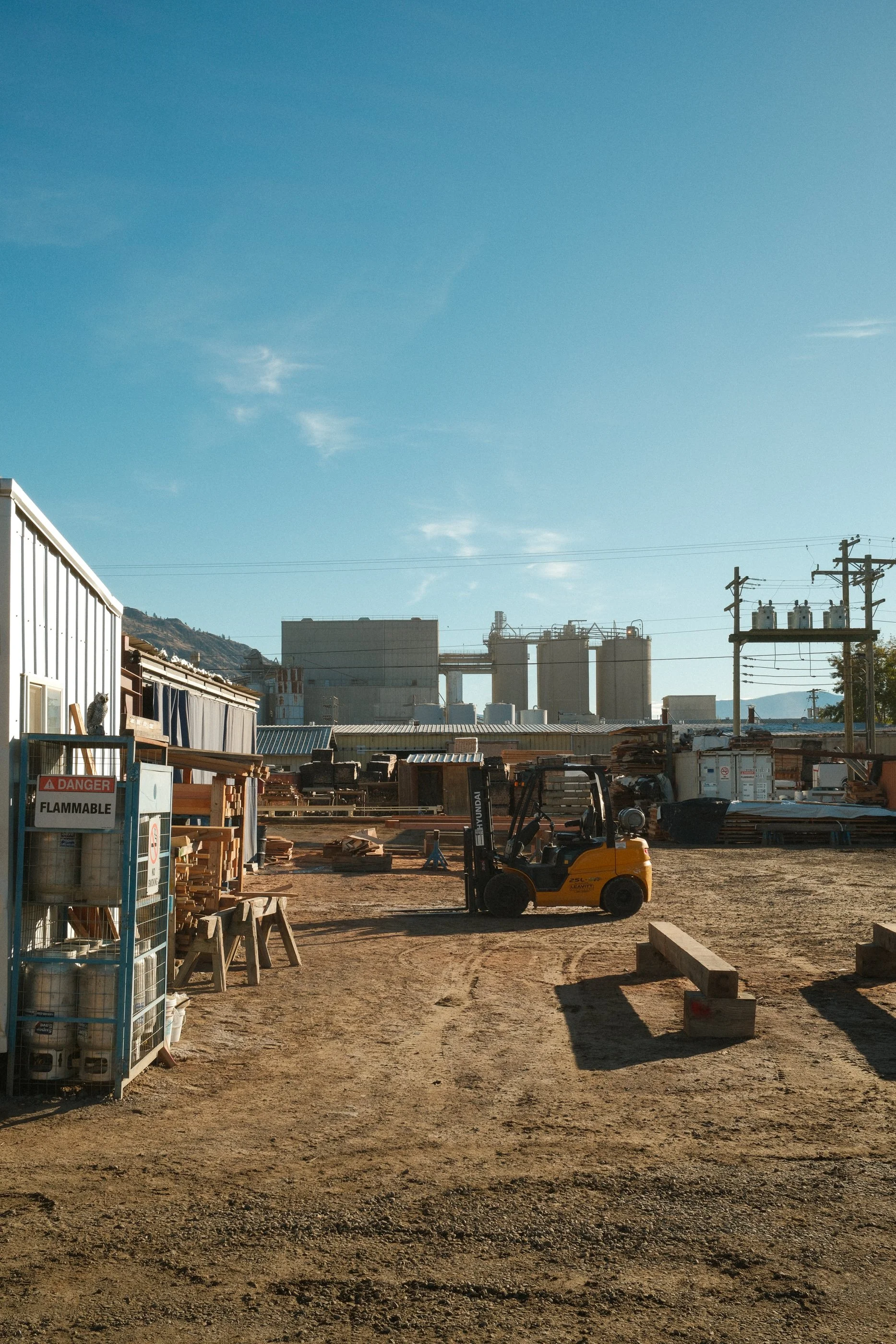 A construction site with a yellow forklift, stacks of wood, and industrial buildings in the background under a clear blue sky.