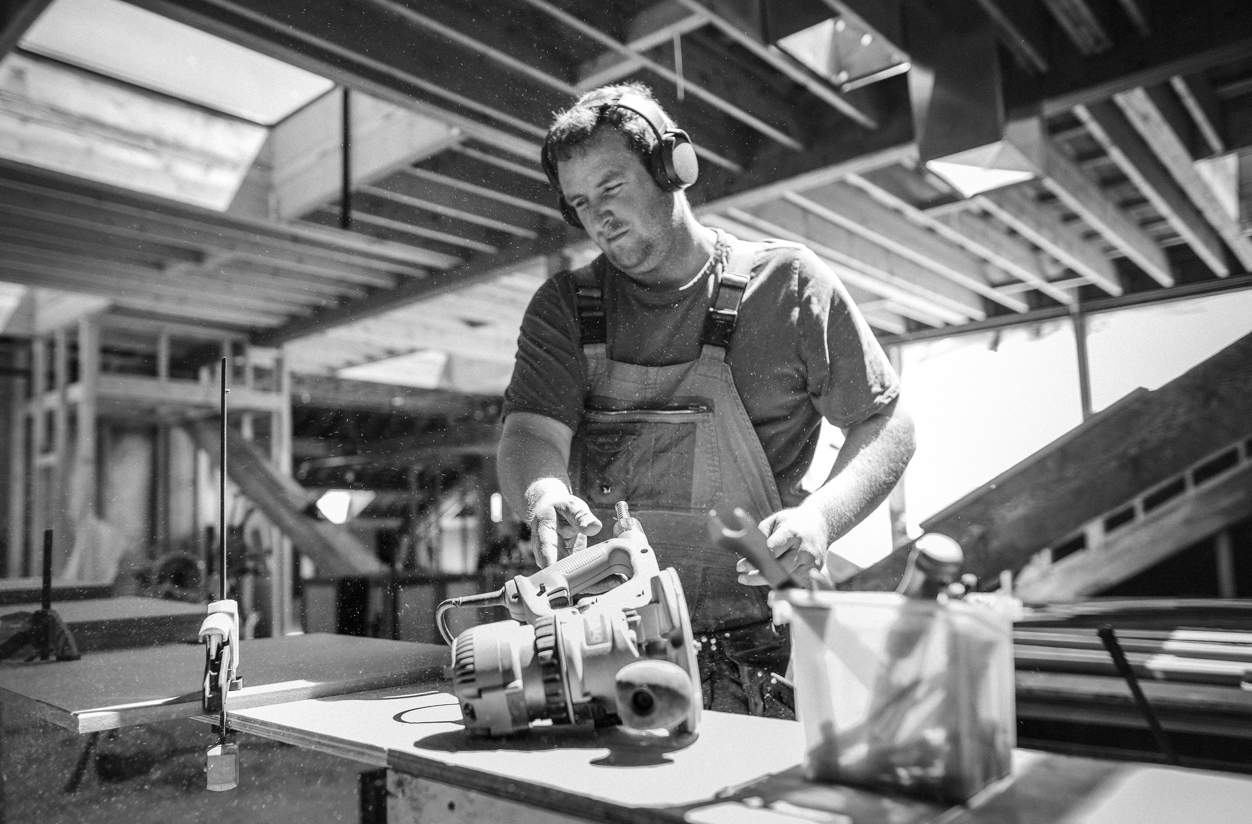A man working with a power saw, wearing overalls and headphones in a construction site with wooden beams.