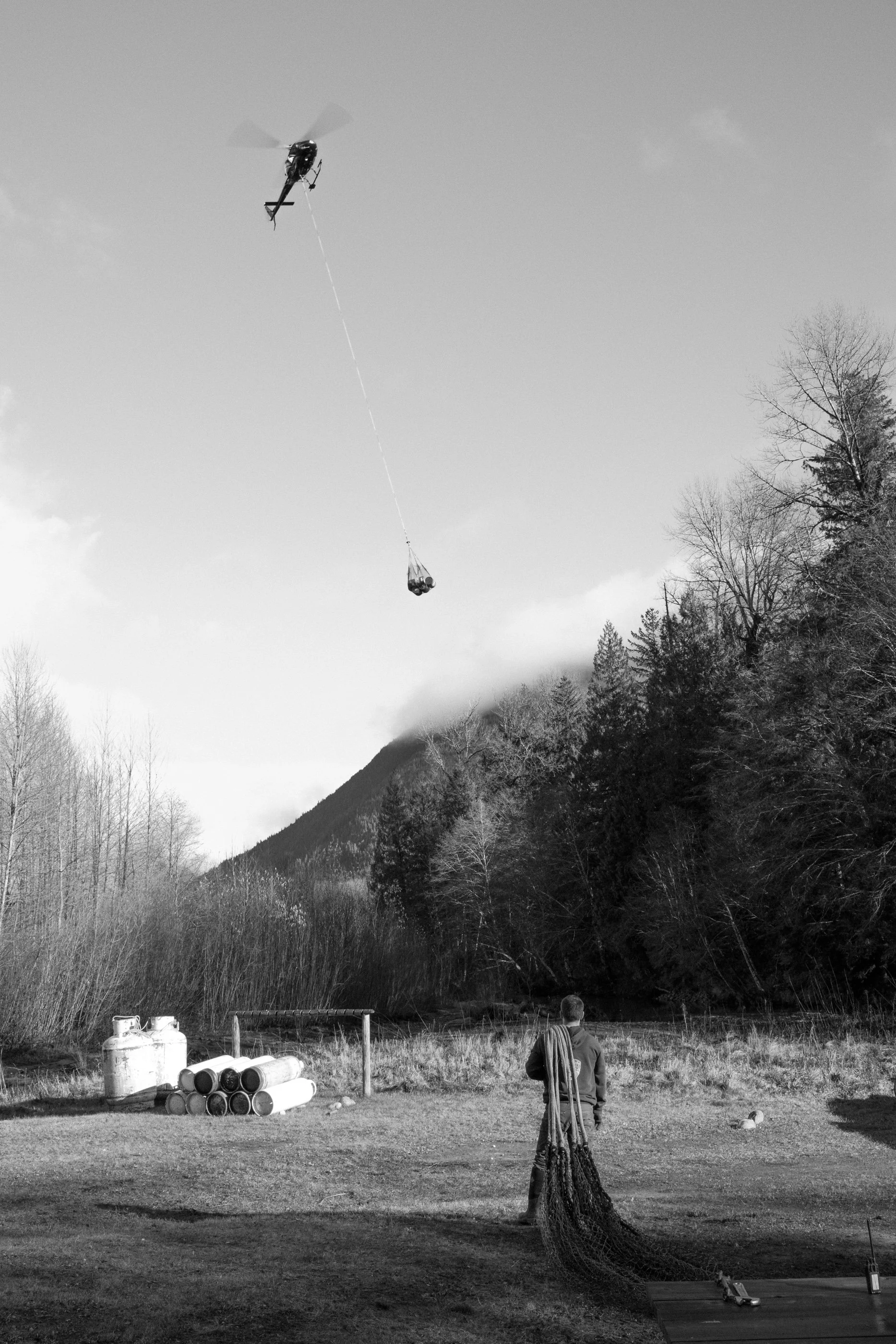 A person standing outdoors holding a fishing net, watching a helicopter flying in the sky pulling a large bag or container, with a mountainous landscape and trees in the background.