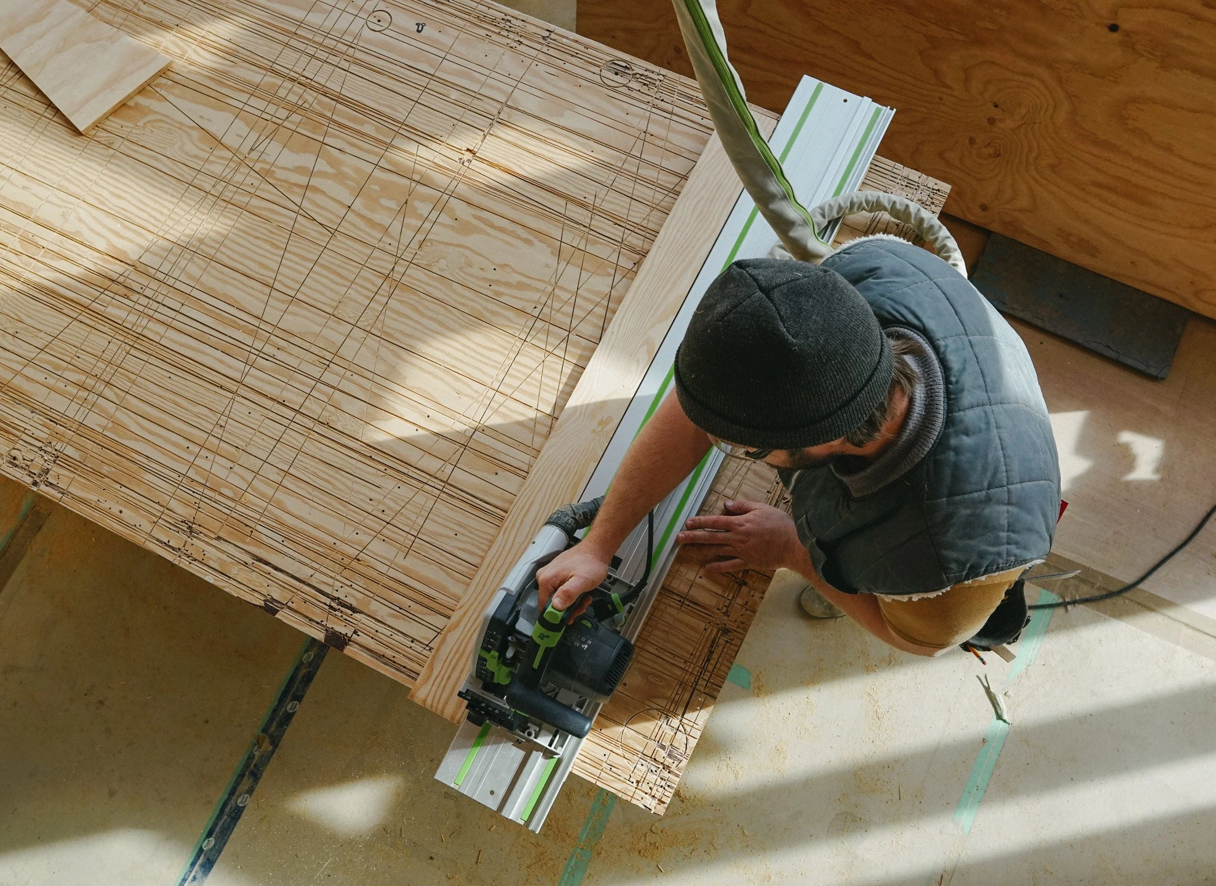 A person wearing a black beanie, glasses, and a blue vest is using a cordless saw to cut a piece of wood on a construction site. The wood appears to be part of a large framework with grid markings. The scene is illuminated by natural light.