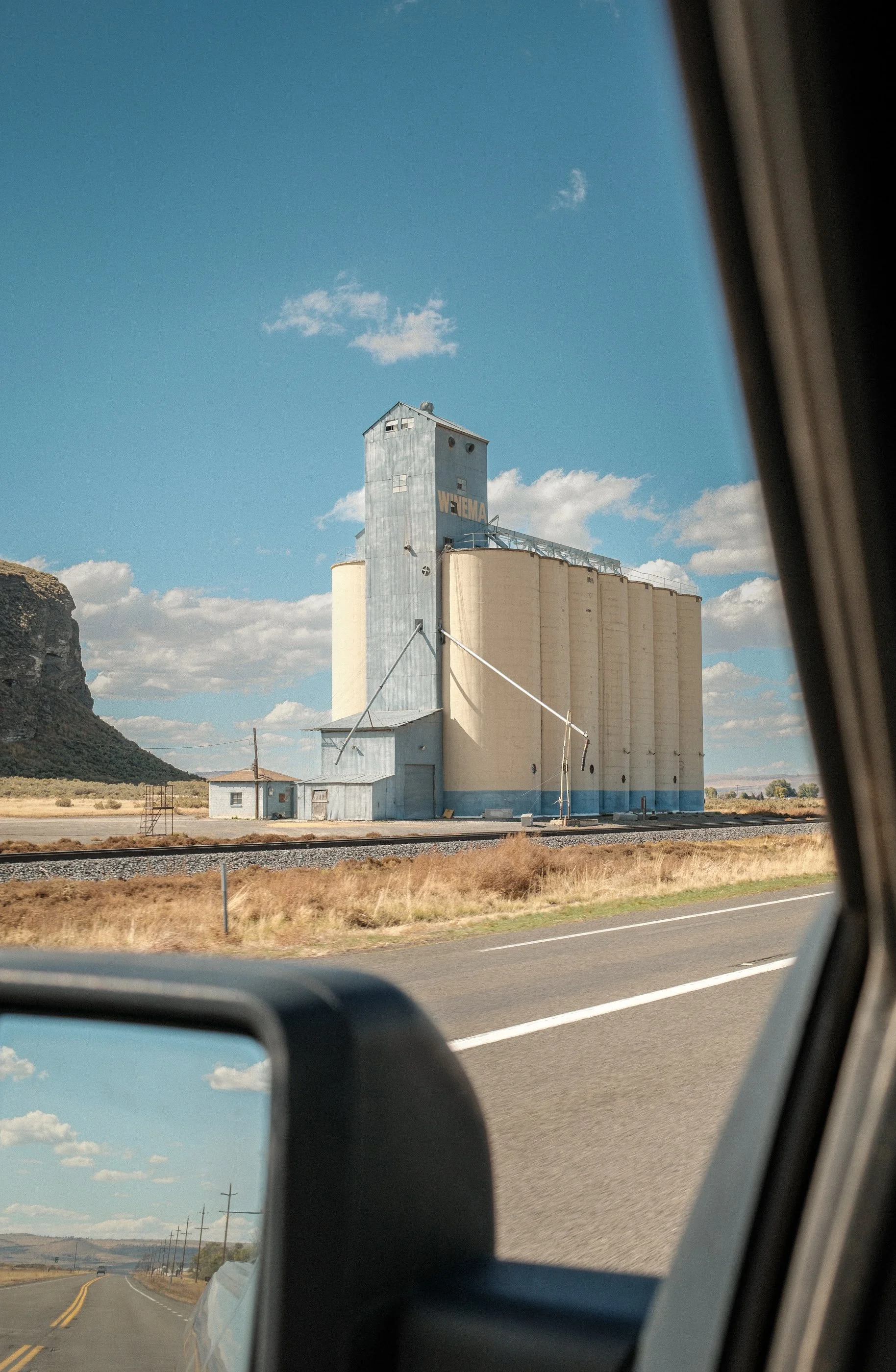View of a large grain elevator with the word 'WEIMER' on it, seen from inside a vehicle on a rural highway with power lines and a mountain in the background.