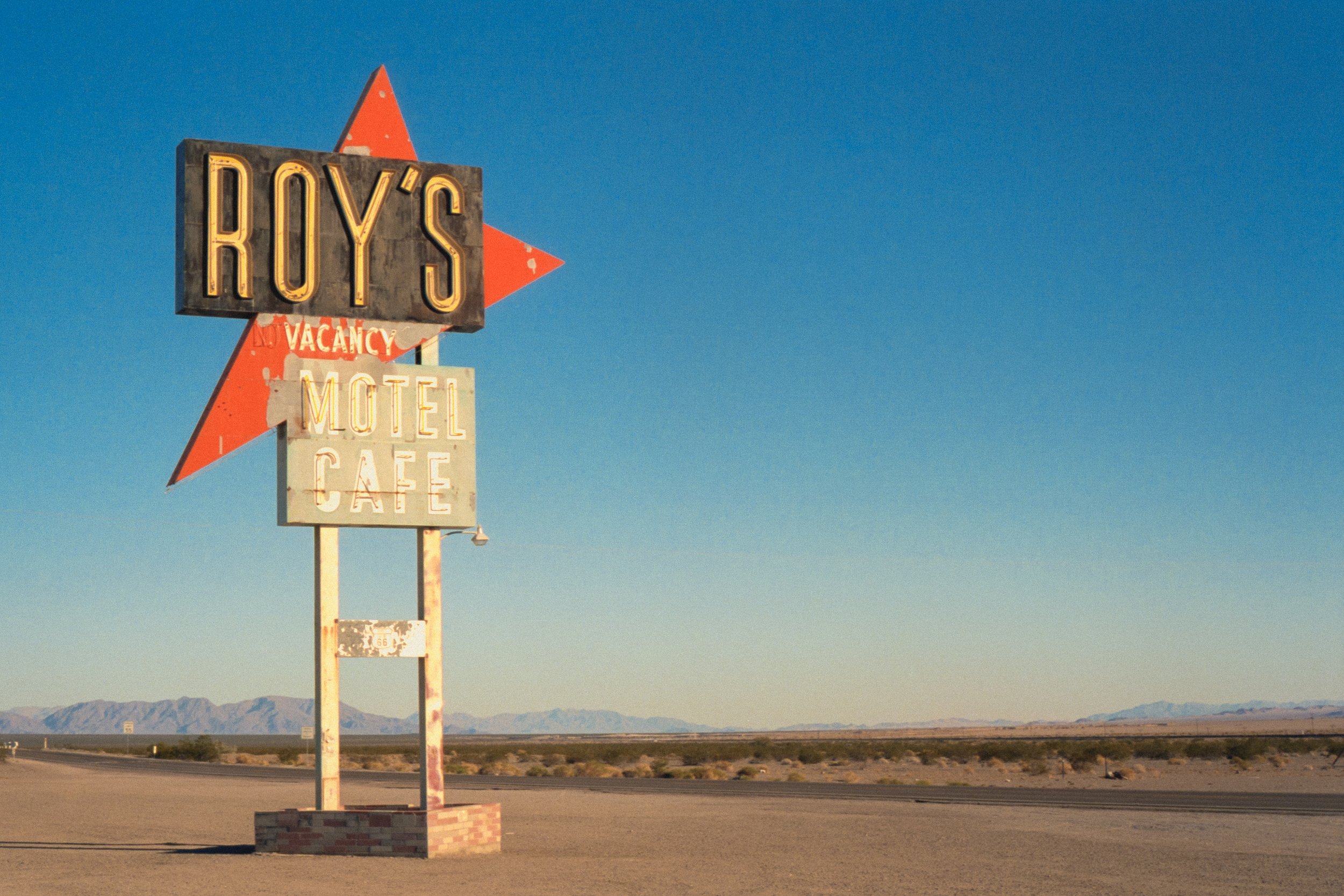 A vintage roadside sign on an empty desert road with mountains in the background, advertising Roy's Motel and Cafe, with parts of the sign showing rust and age.