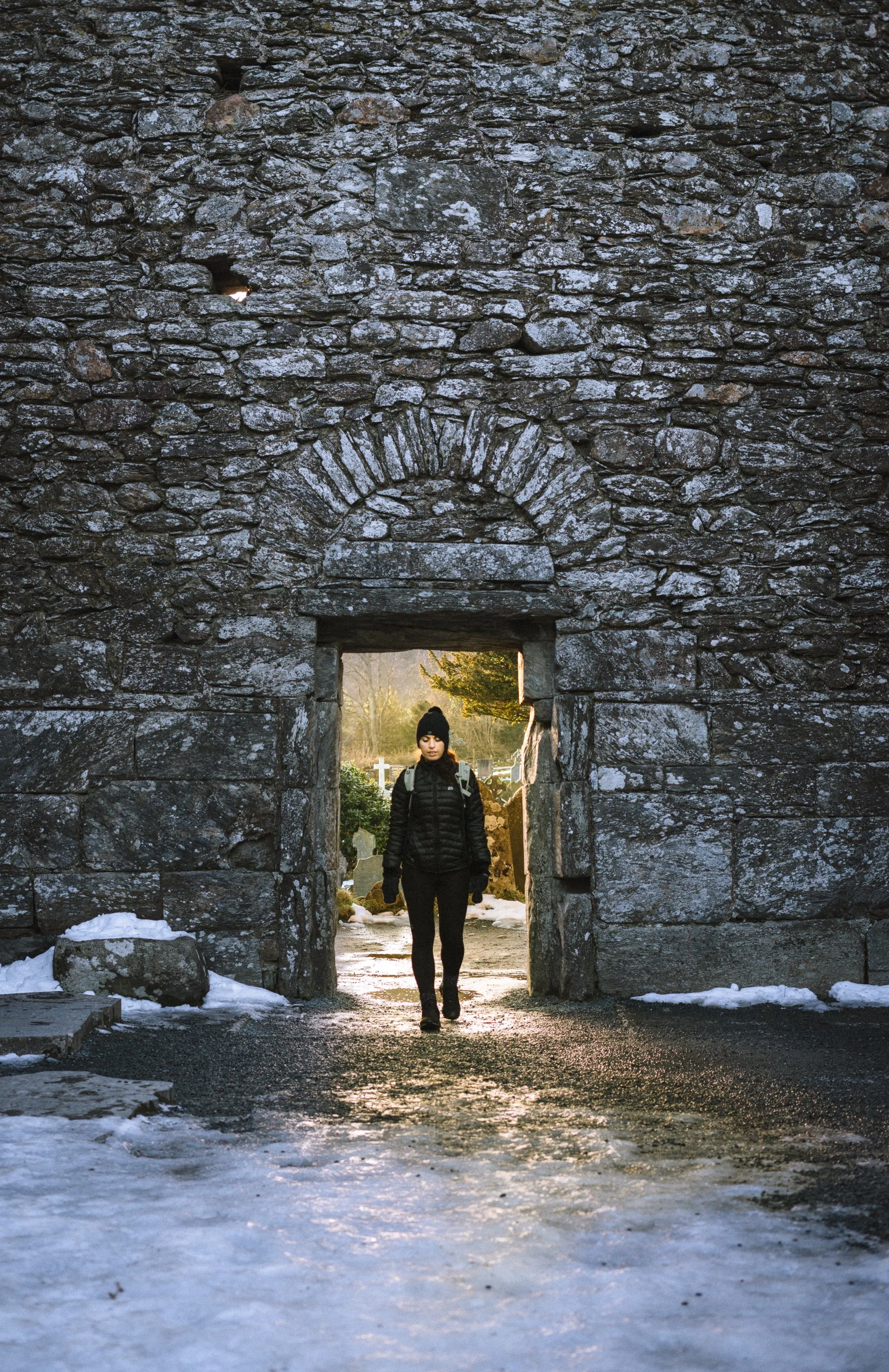 A person wearing a black jacket, black pants, and a black beanie walking through an arched stone gateway on a cold day, with snow on the ground and trees in the background.