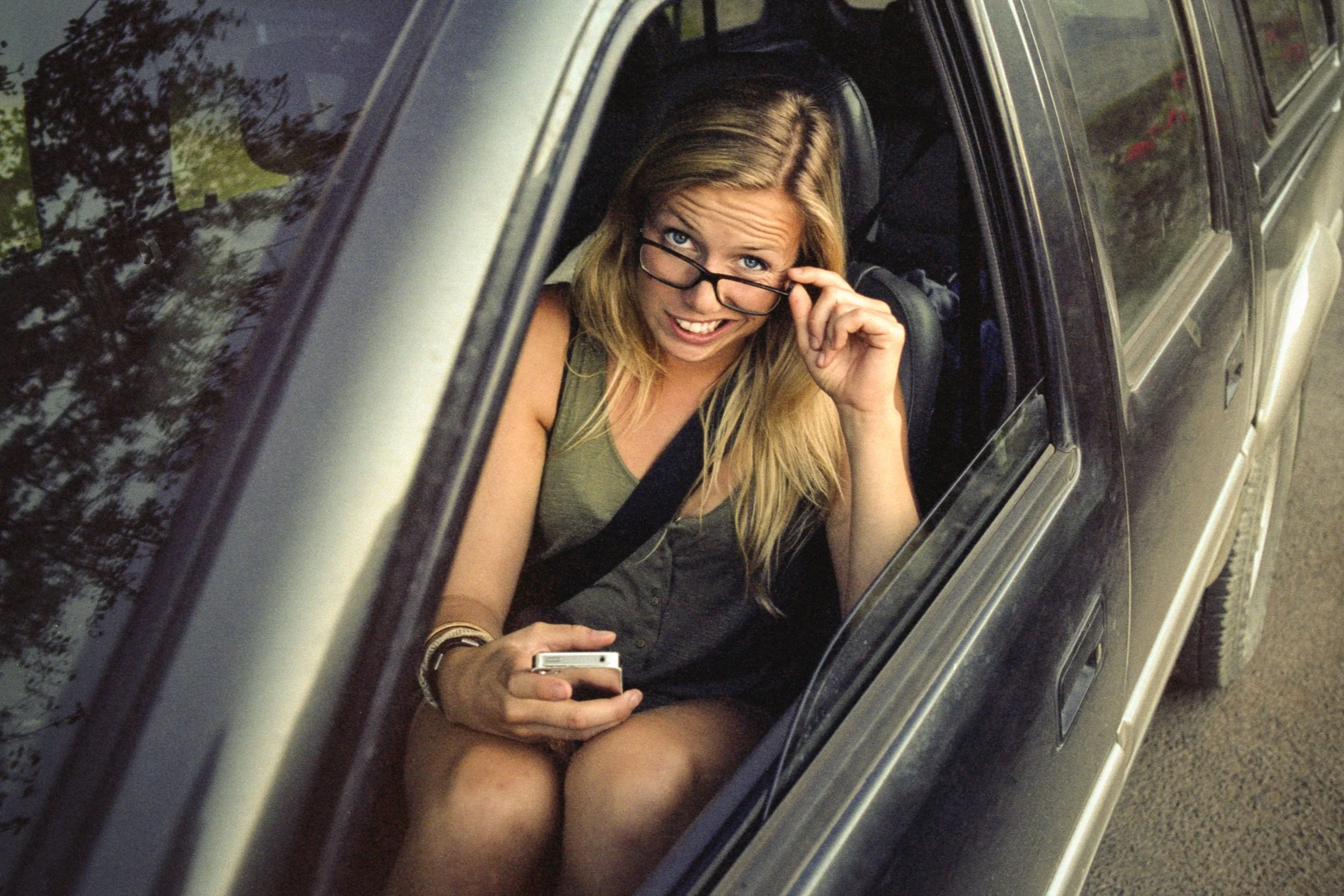 A woman sitting in the front passenger seat of a car, looking up at the camera and adjusting her glasses while holding a smartphone.