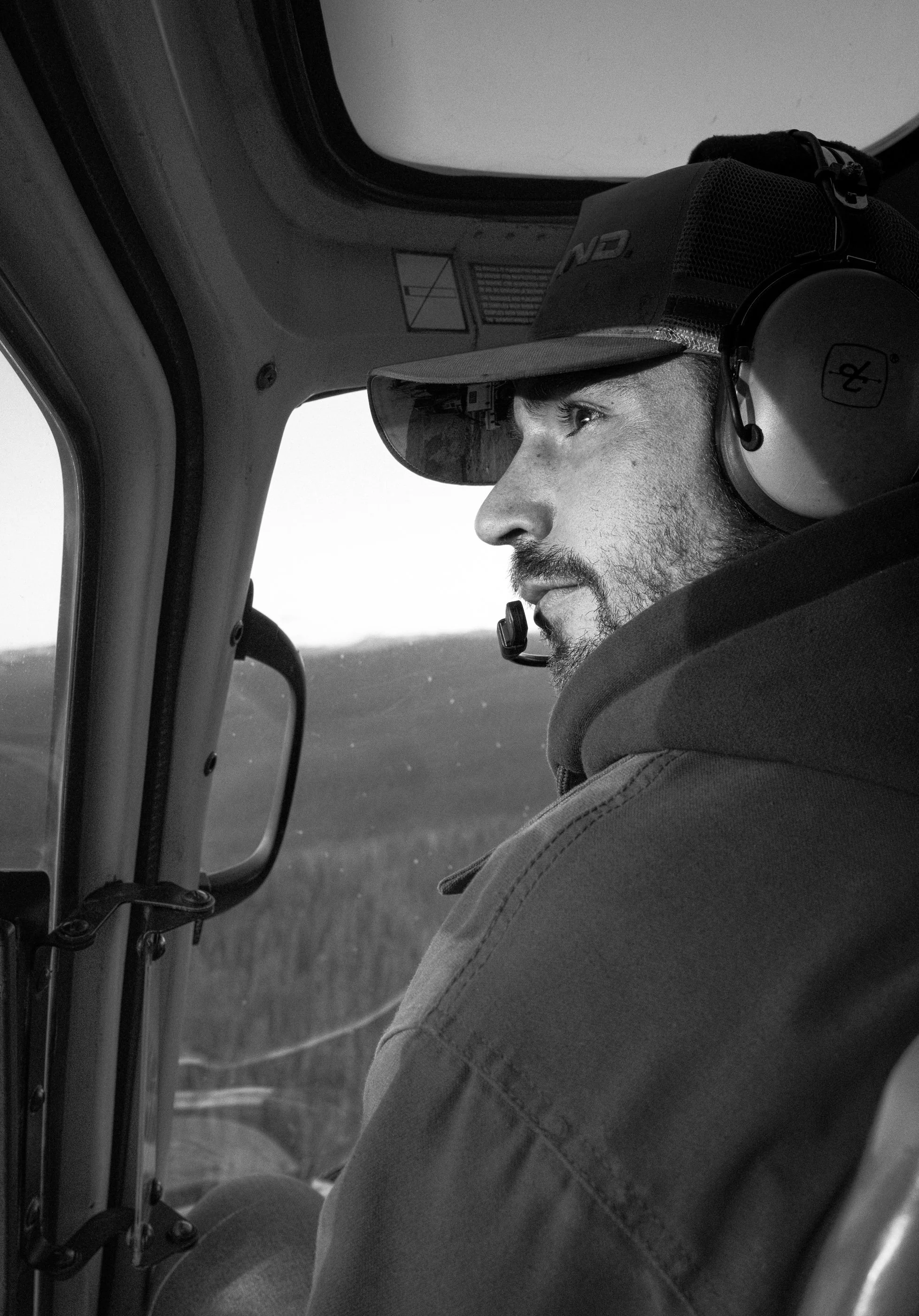 A man with a beard wearing a cap and headphones, sitting in the cockpit of an aircraft, looking outside through the window.