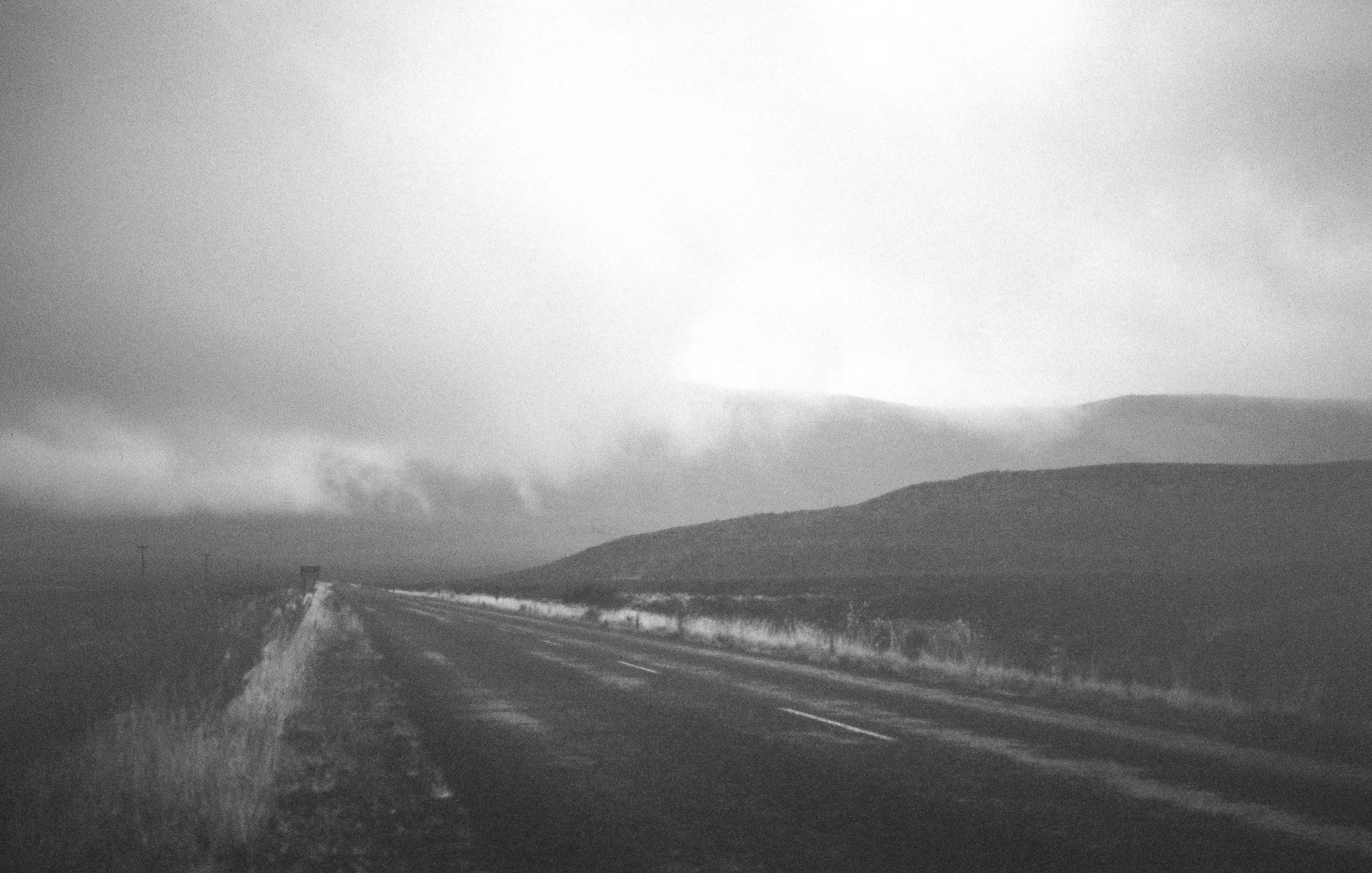 Black and white photo of a deserted road stretching into the distance in a rural area with hills on the horizon and cloudy sky overhead.