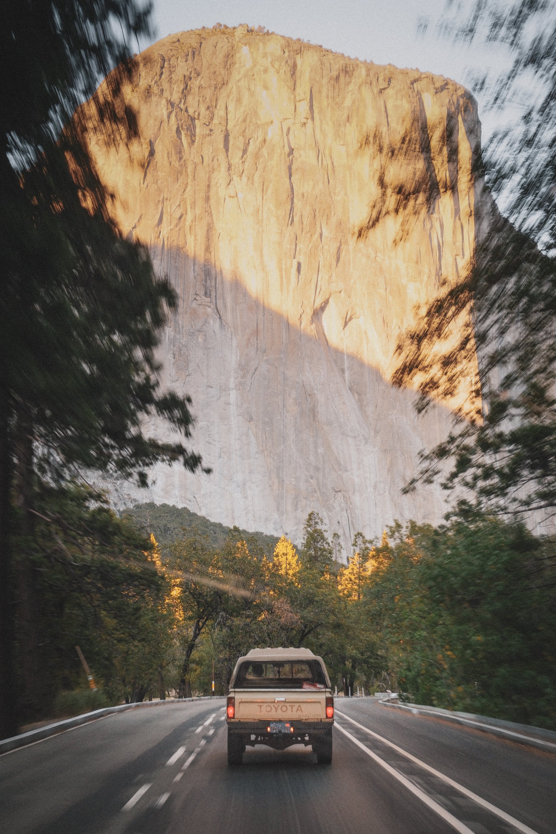 A pickup truck driving on a winding road through a forest with trees showing fall colors, with a large rock formation or mountain in the background.
