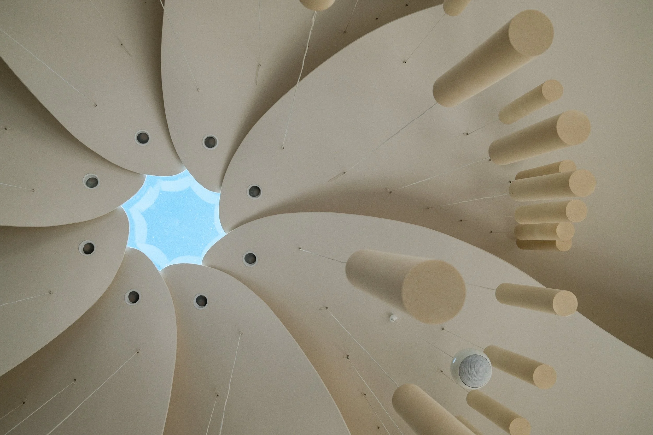 Looking up at a modern chandelier with multiple cylindrical and rounded shapes hanging from thin wires, surrounding a light blue sky viewed through a circular opening.