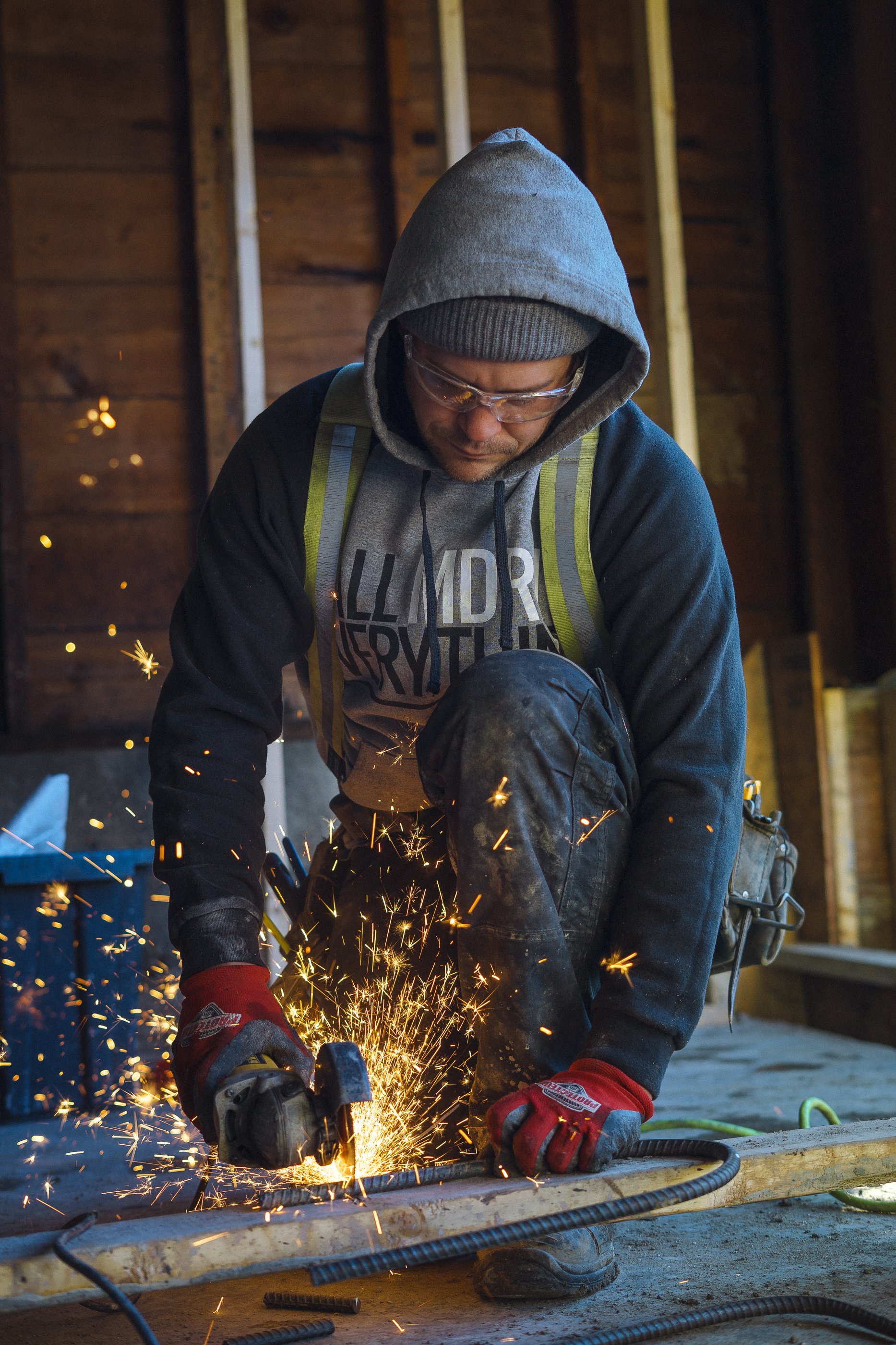 A man in a gray hoodie, safety glasses, and red gloves is welding metal inside a wooden workshop, with sparks flying as he works.