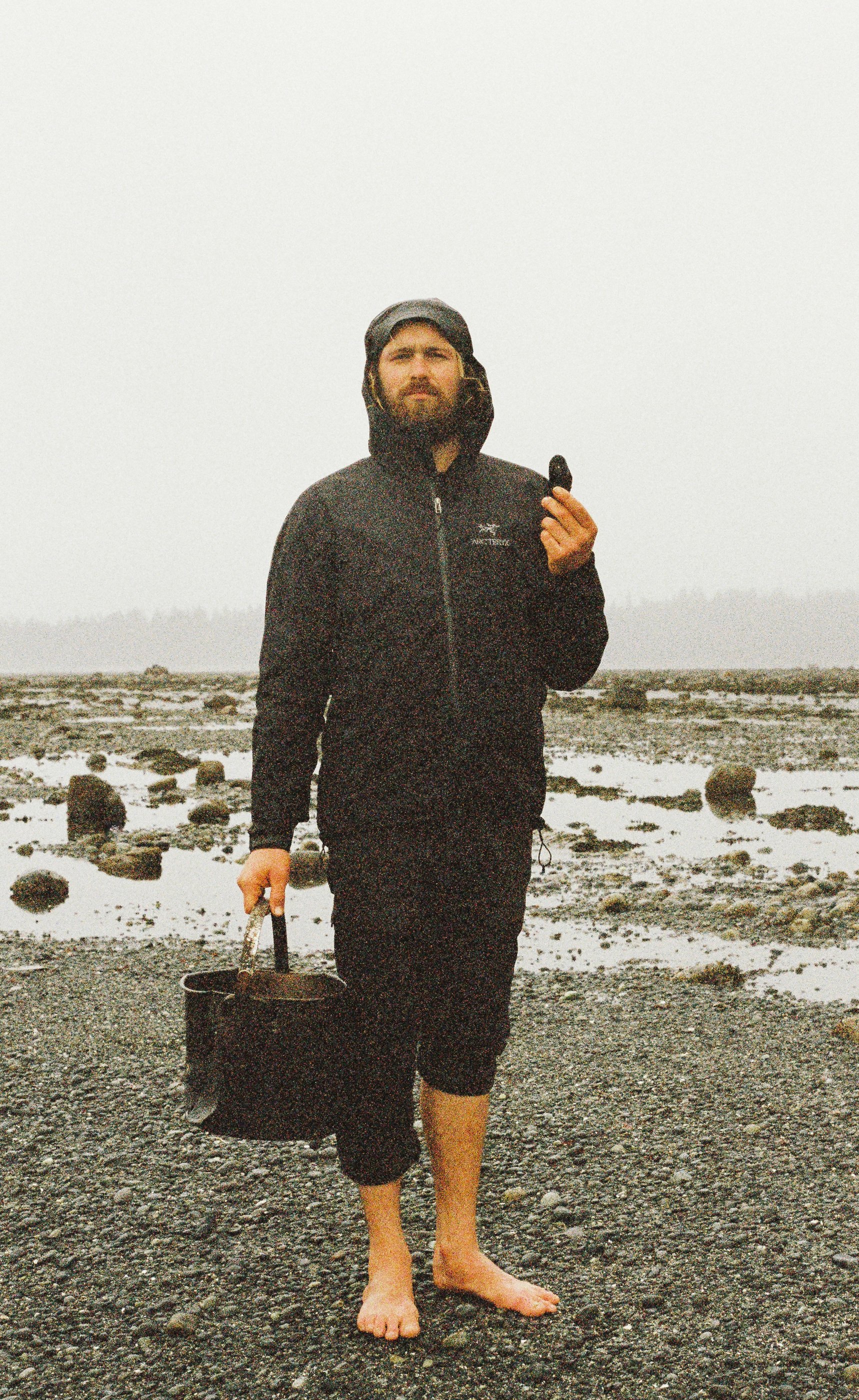 A man dressed in black jacket and pants, barefoot, standing on rocky and wet terrain, holding a bucket in one hand and a small object in the other, with a foggy, overcast background.