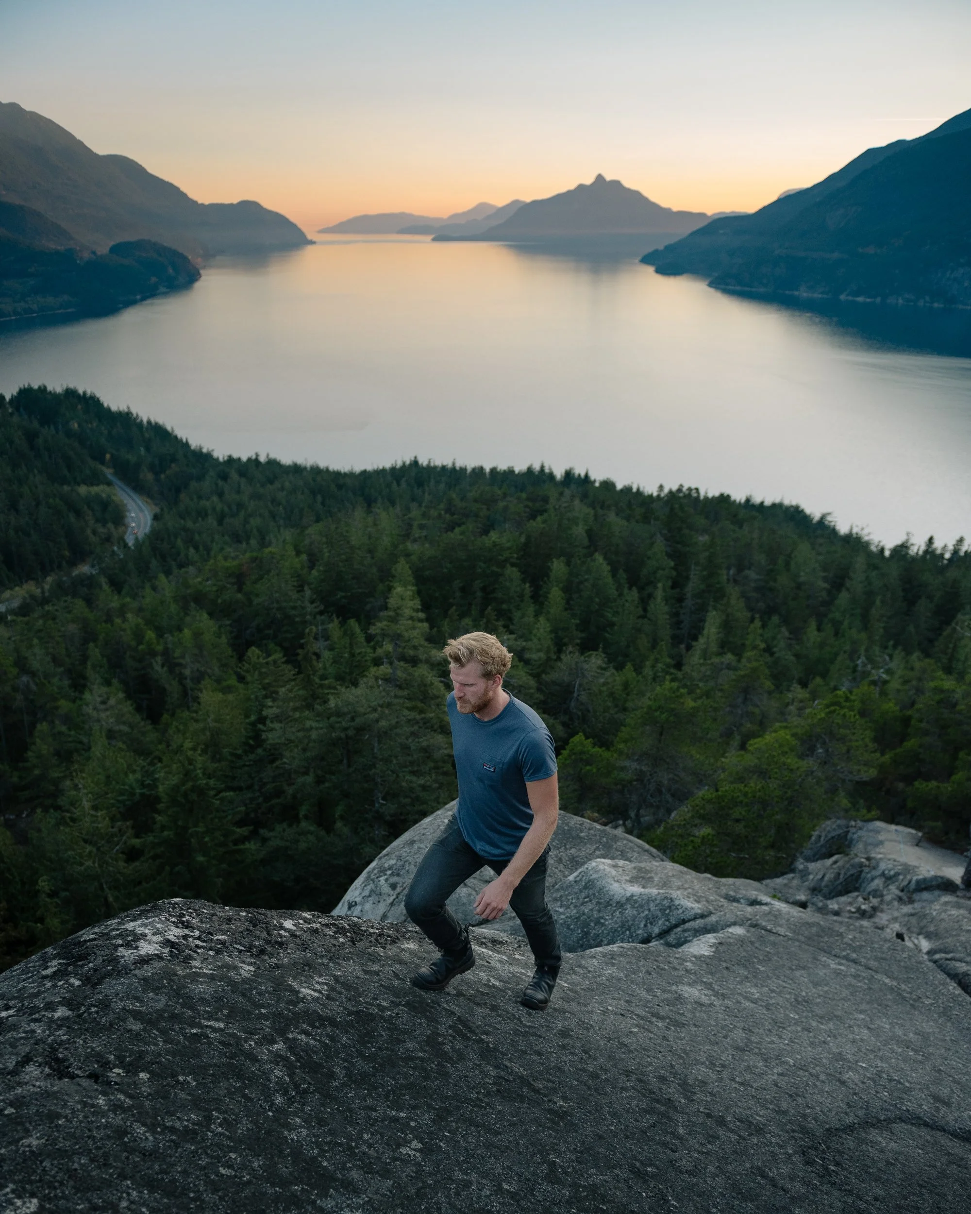 A man climbing on a large rock formation with a scenic lake, forest, and mountains in the background at sunset.