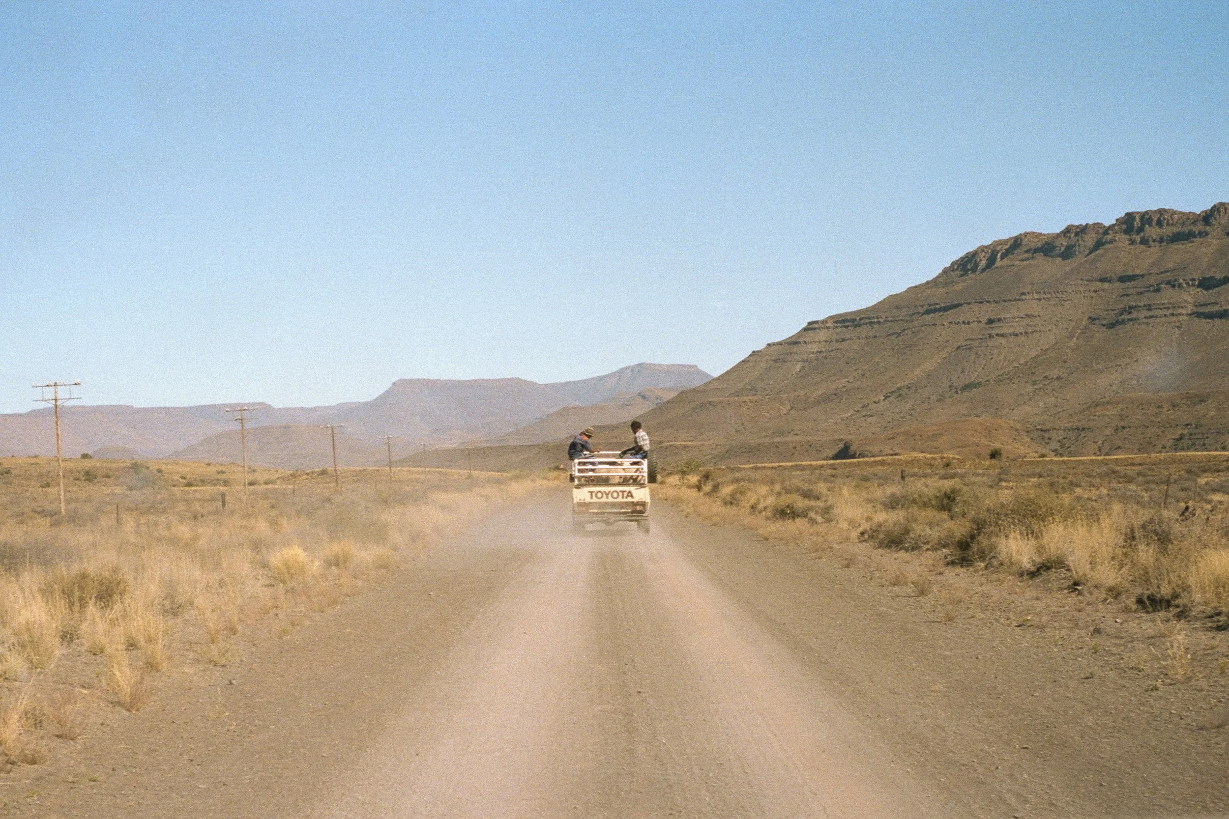 A pickup truck driving on a dirt road in a desert landscape with mountains in the background, two people sitting in the truck's bed.