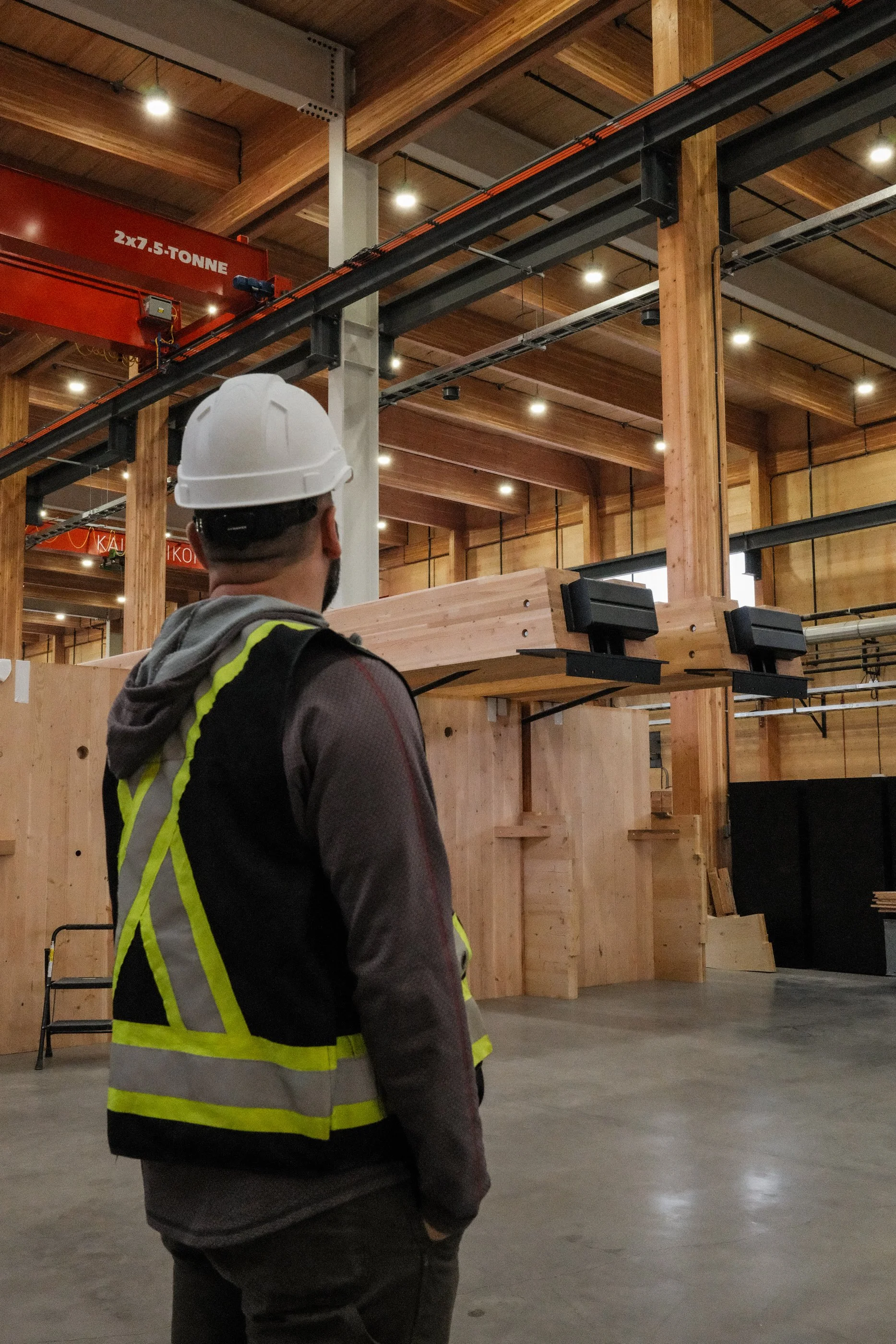 A construction worker wearing a white safety helmet and a black vest with yellow reflective stripes stands inside a wooden building under construction. The building features a high ceiling with exposed wooden beams and a red overhead crane labeled "2