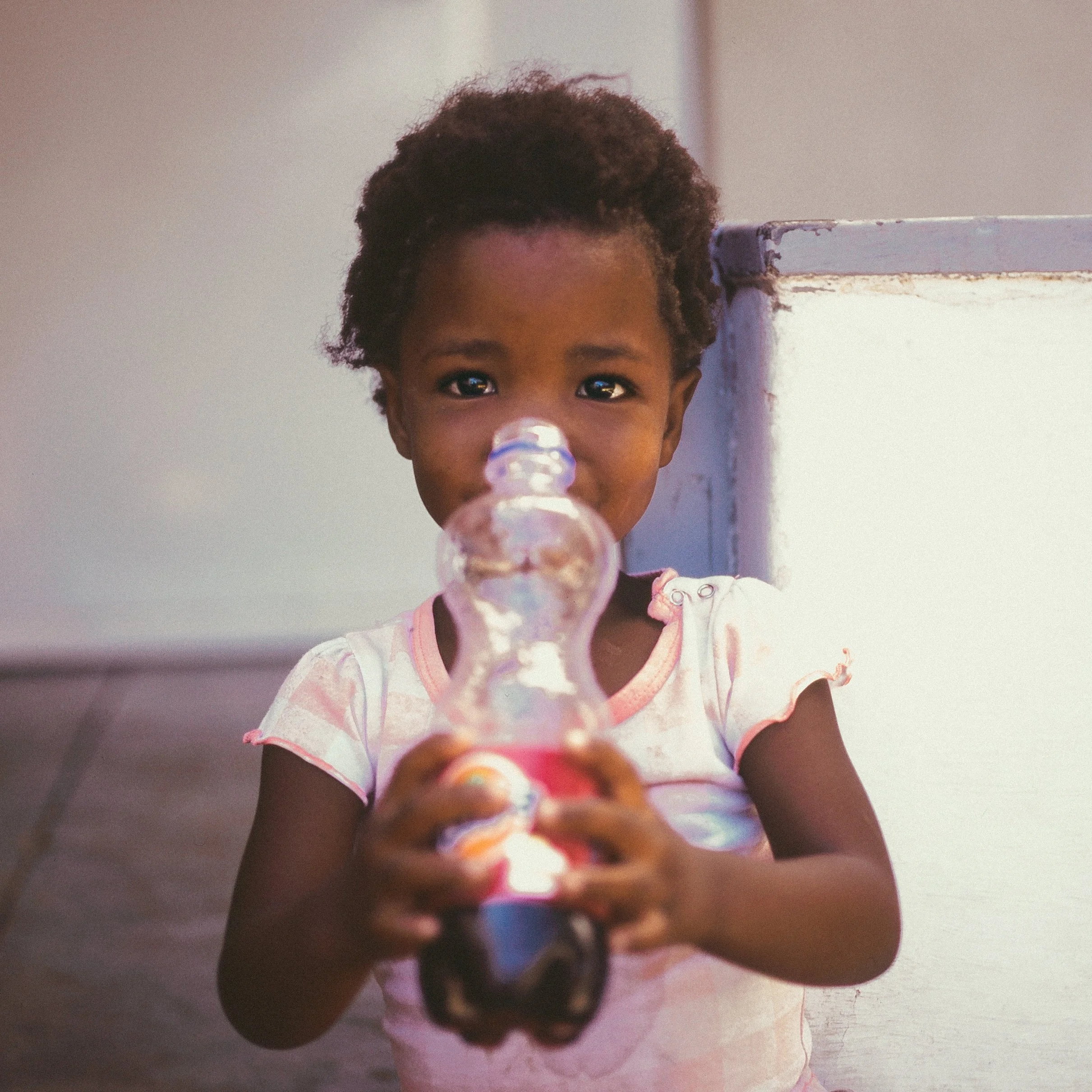 A young girl with dark curly hair, wearing a light pink and white shirt, holding a plastic bottle and looking directly at the camera.