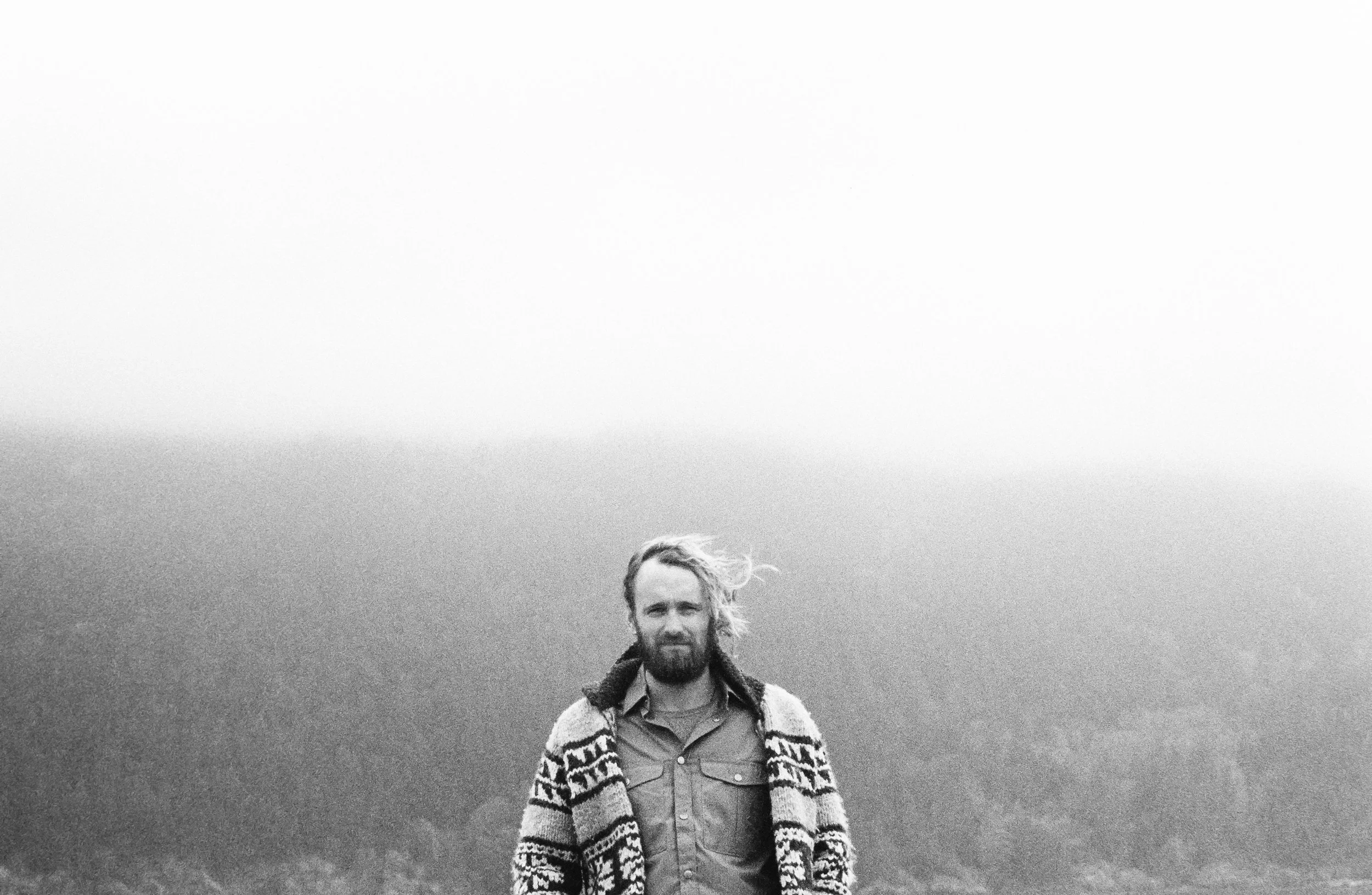Black and white photo of a bearded man with long hair standing outdoors with mountains and sky in the background, wearing a patterned sweater and shirt.