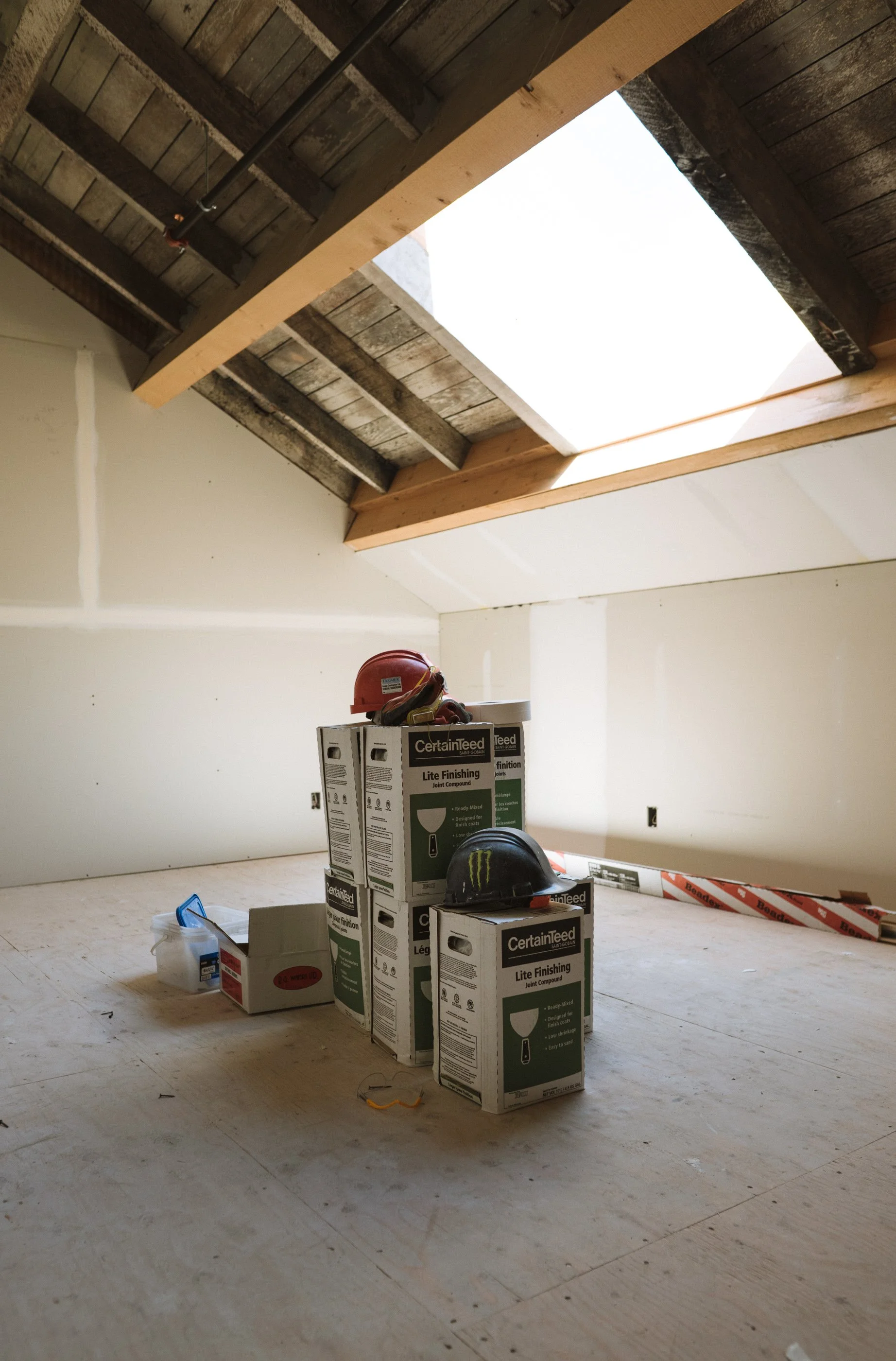 Room under construction with boxes of CertainTeed Lite Finishing joint compound and protective helmets on the floor, and an open skylight in the ceiling with exposed beams.