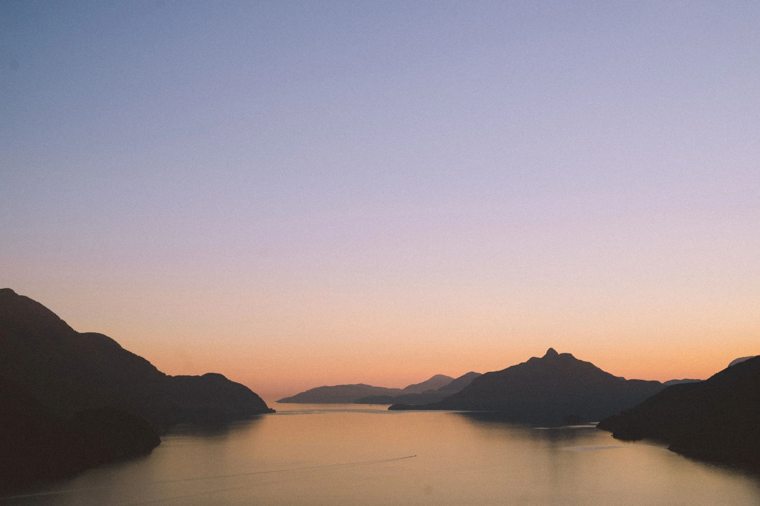 Peaceful view of a river or lake at sunset, with mountains in the distance and a clear sky with pastel colors.