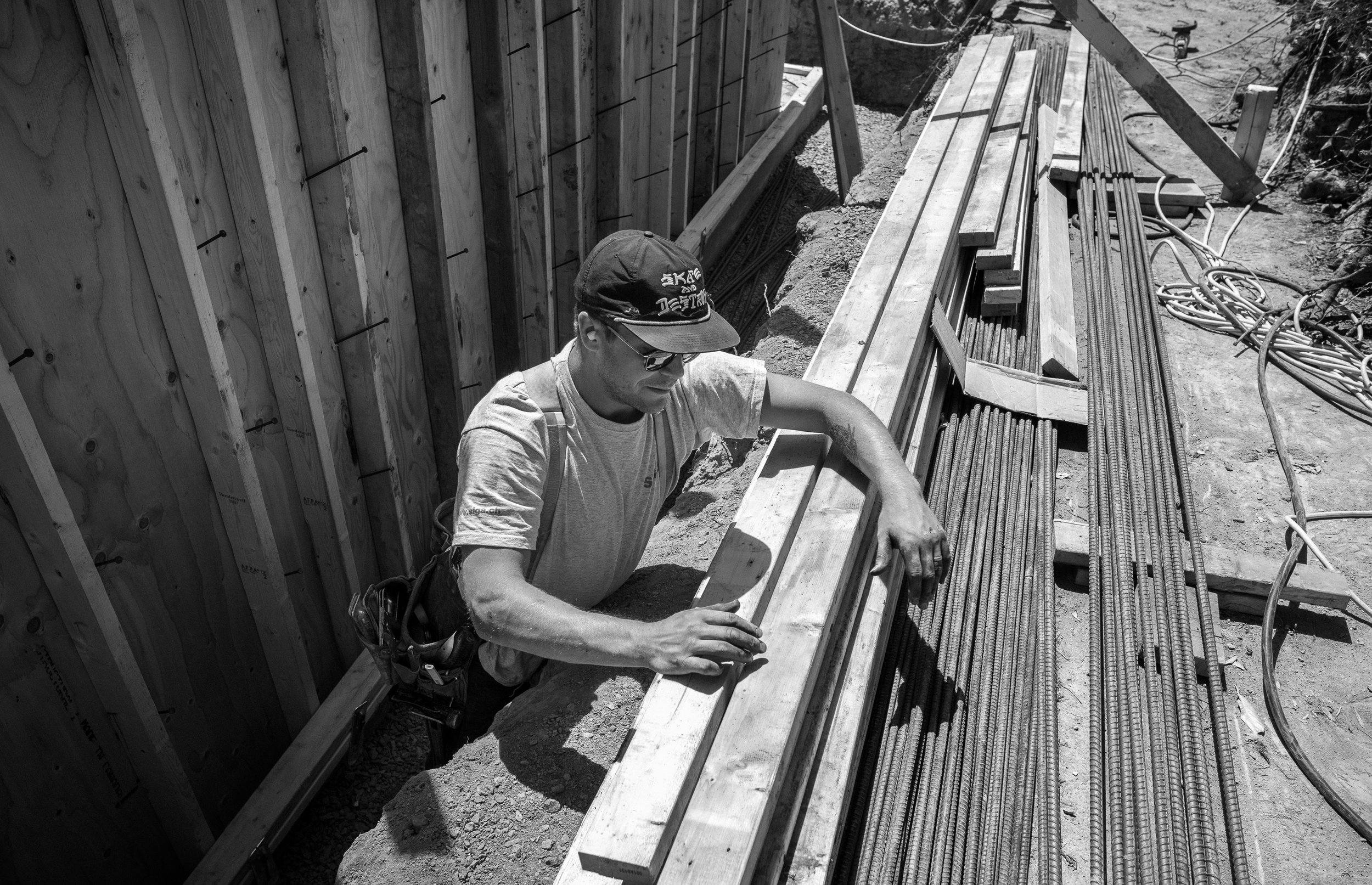 A construction worker leaning through an opening in a wall, working with a wooden plank at a construction site with steel rebar and construction tools laying on the ground.