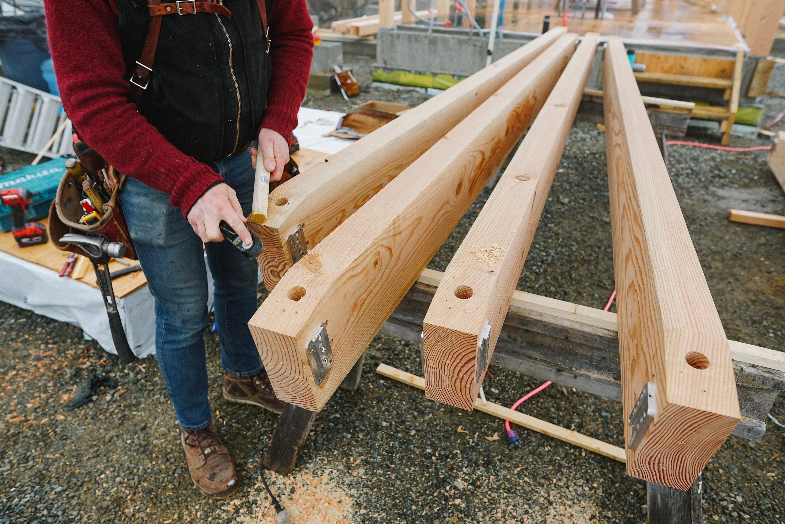 A person working on a woodworking project at a construction site, assembling long wooden beams with metal brackets.