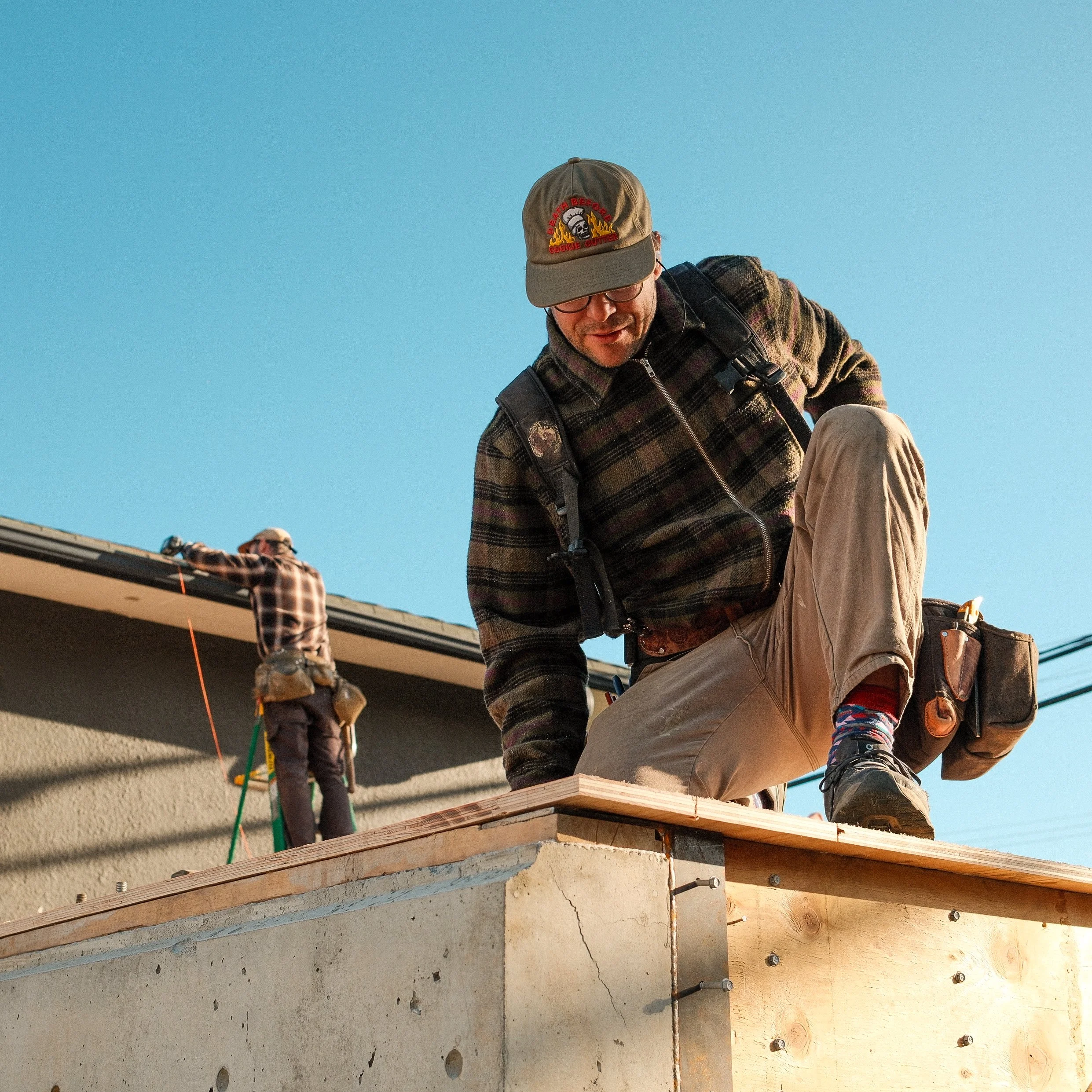Two construction workers, one close-up kneeling on a roof, and the other in the background standing and working on the roof, both wearing plaid shirts, hats, and tool belts. Clear blue sky.