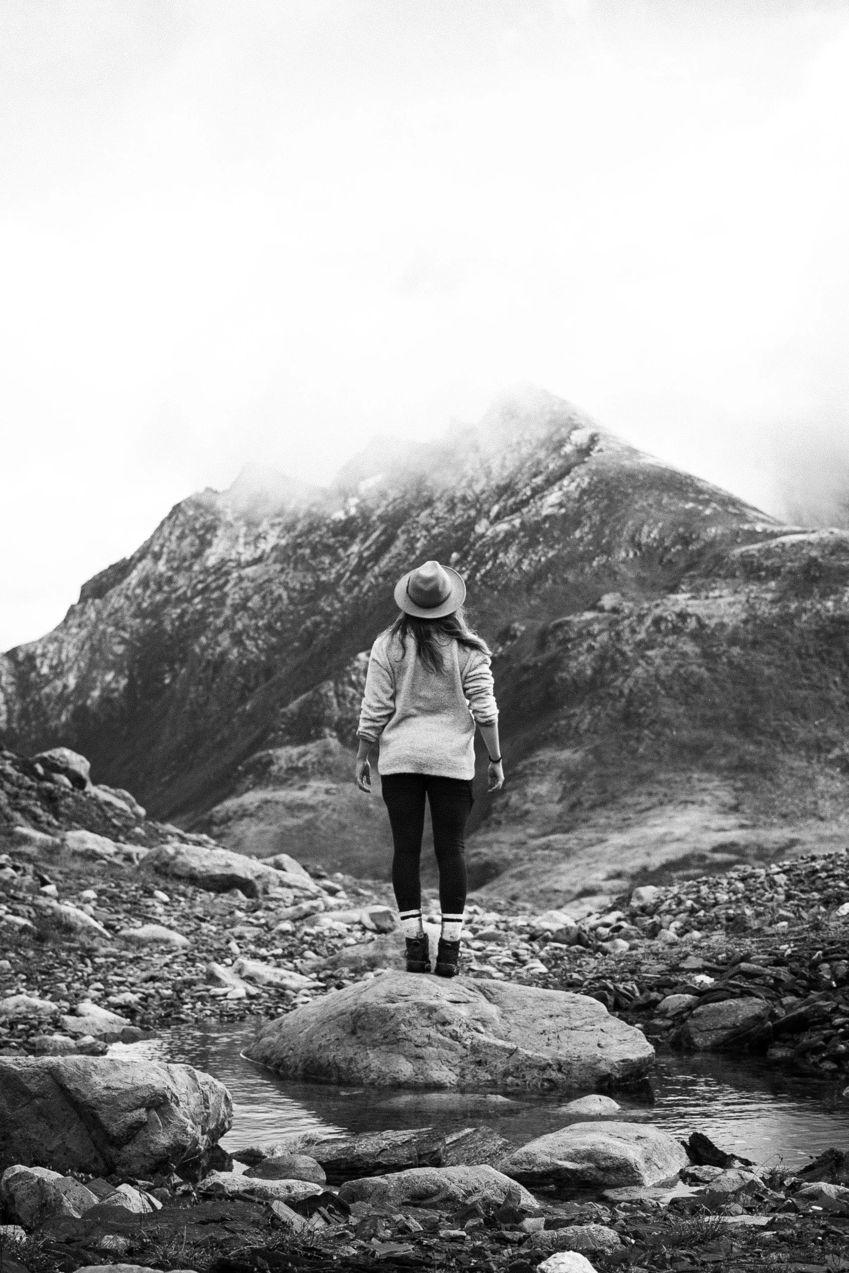 A woman standing on a rock in a stream, looking at a mountain in a cloudy landscape, in black and white.