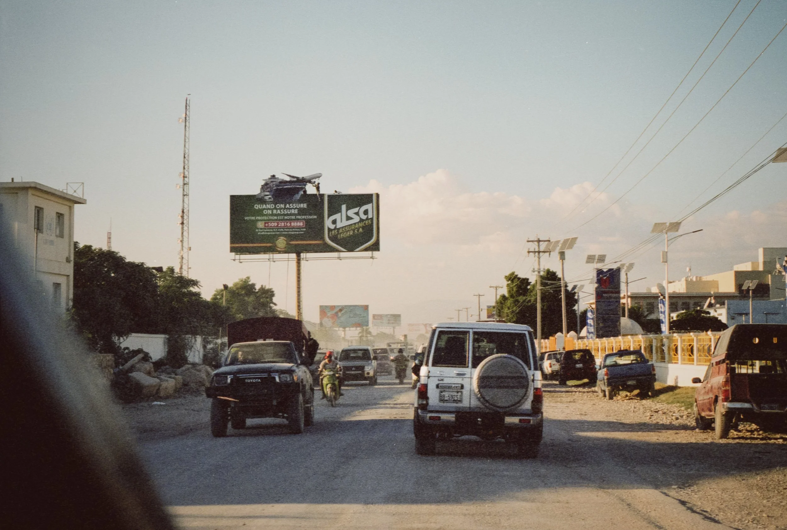 A busy street scene with several cars driving, including a white SUV in the foreground and a dark truck and motorcycle among other vehicles. There are utility poles with solar panels and billboards, one featuring a helicopter and insurance promotion.