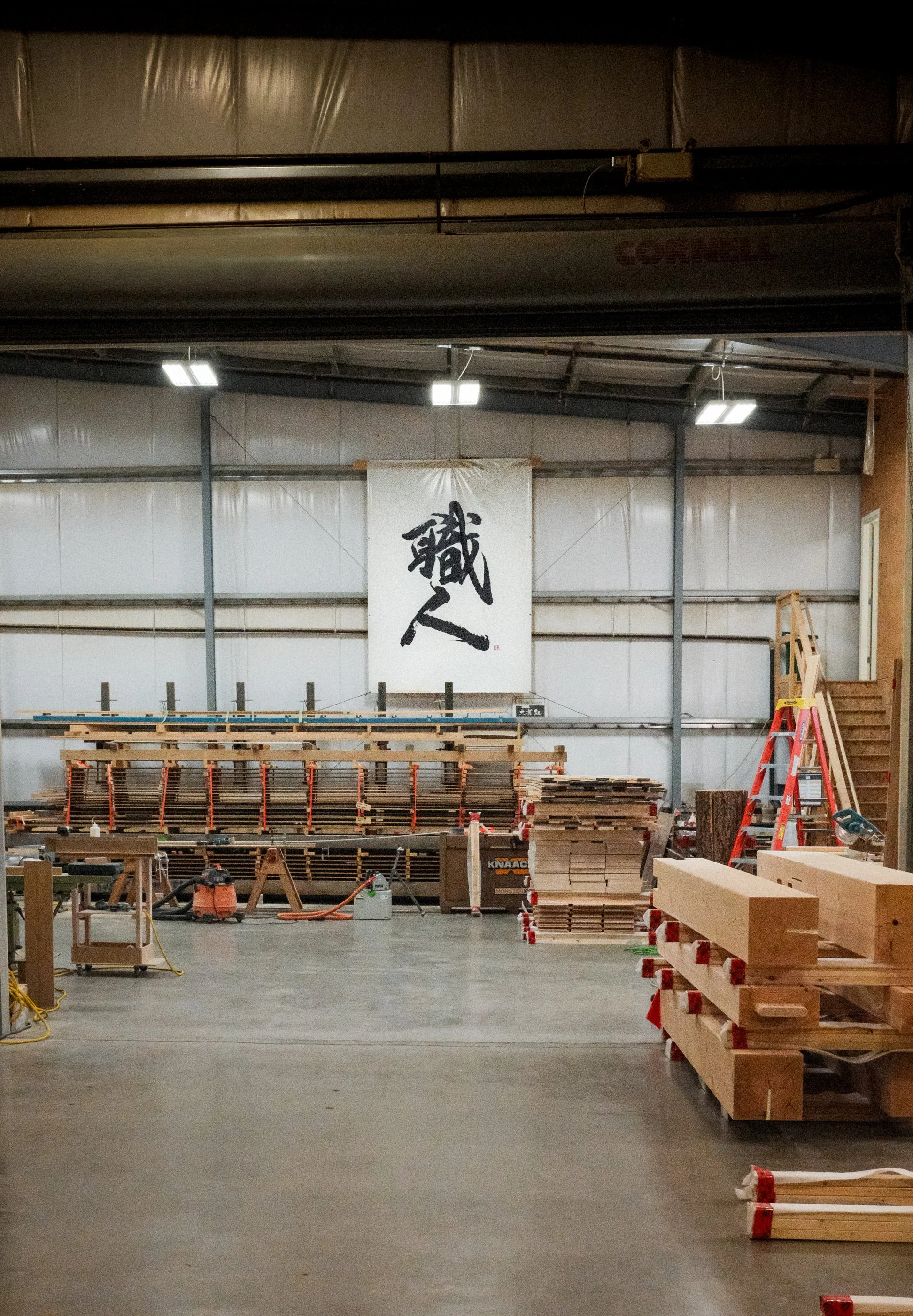 Interior view of a woodworking workshop with stacked lumber, a ladder, and tools, featuring a large Japanese calligraphy banner hanging on the wall.