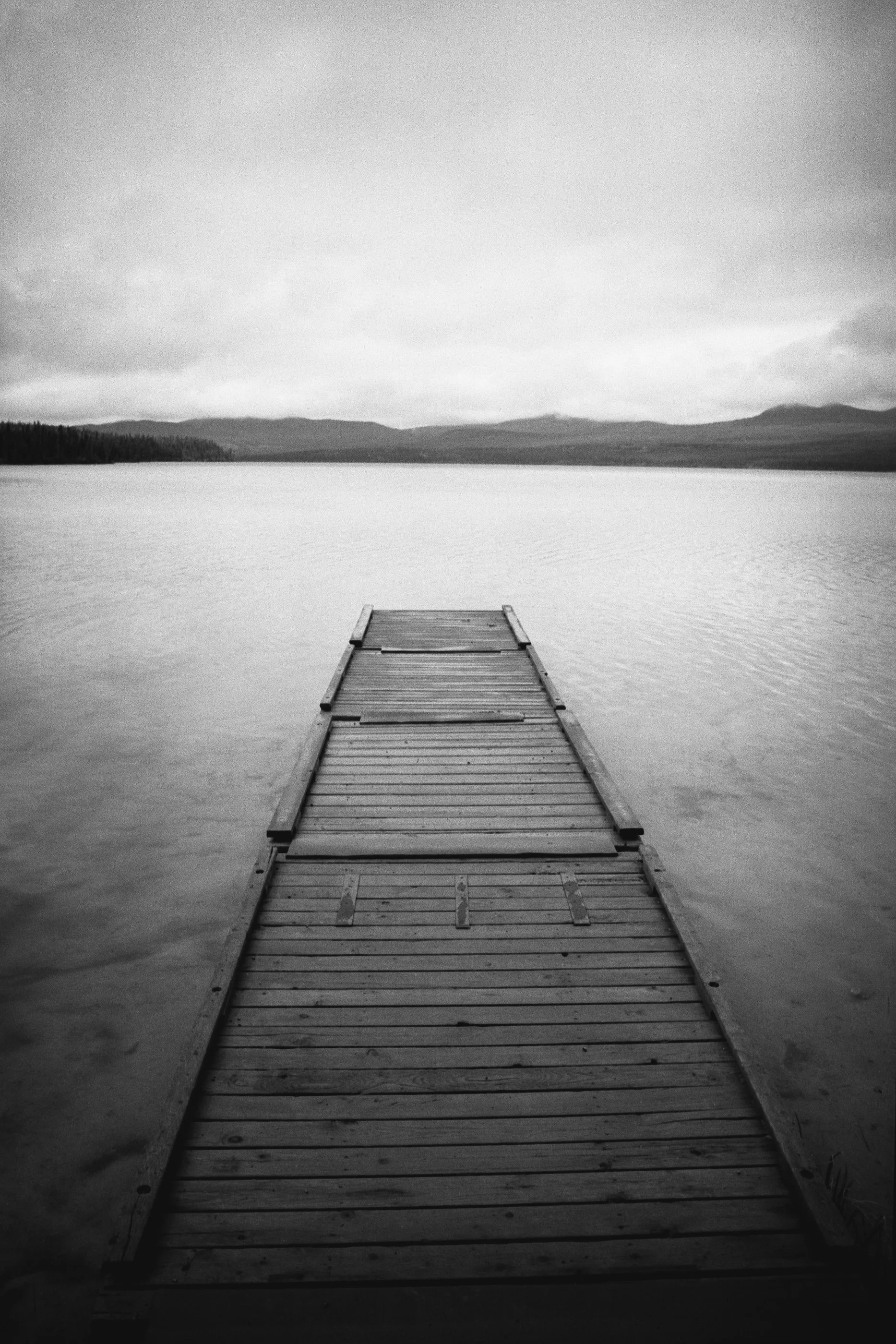 A black and white photo of a wooden dock extending over calm water, with distant hills and a cloudy sky in the background.
