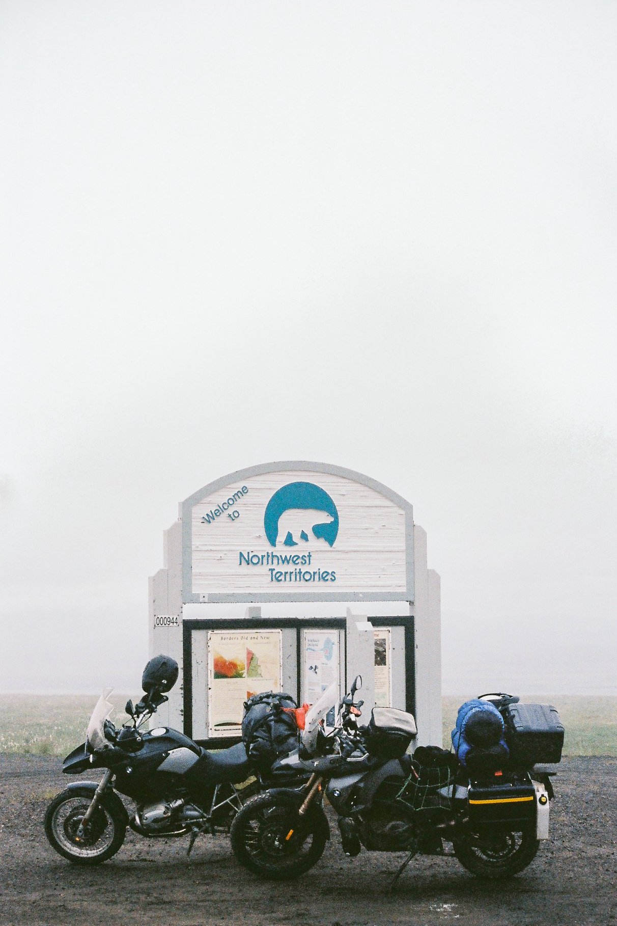 Two motorcycles parked in front of a welcome sign for the Northwest Territories, with a misty background.