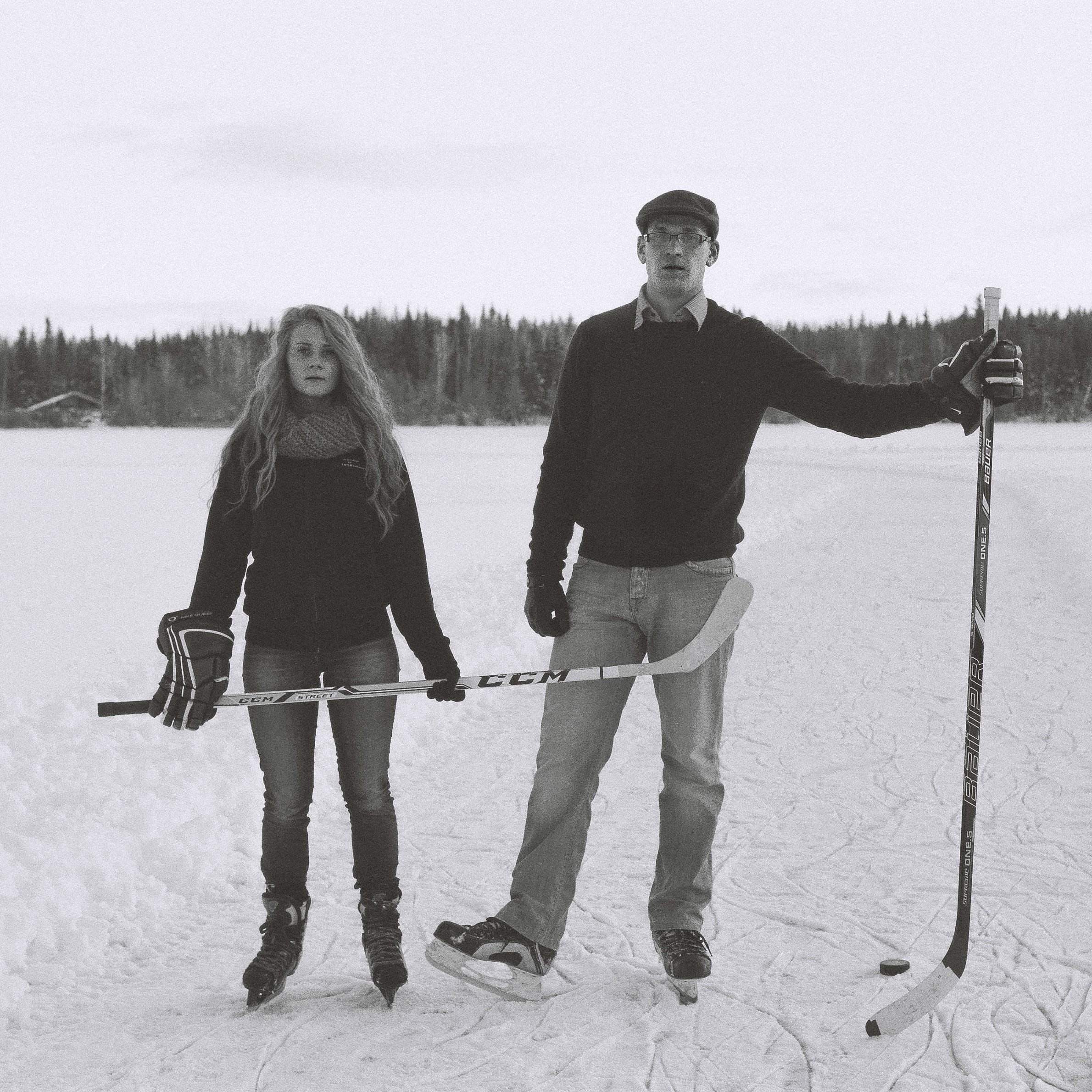 A black-and-white photo of a young woman and a man standing on a snow-covered ice rink outdoors, holding hockey sticks and wearing ice skates.