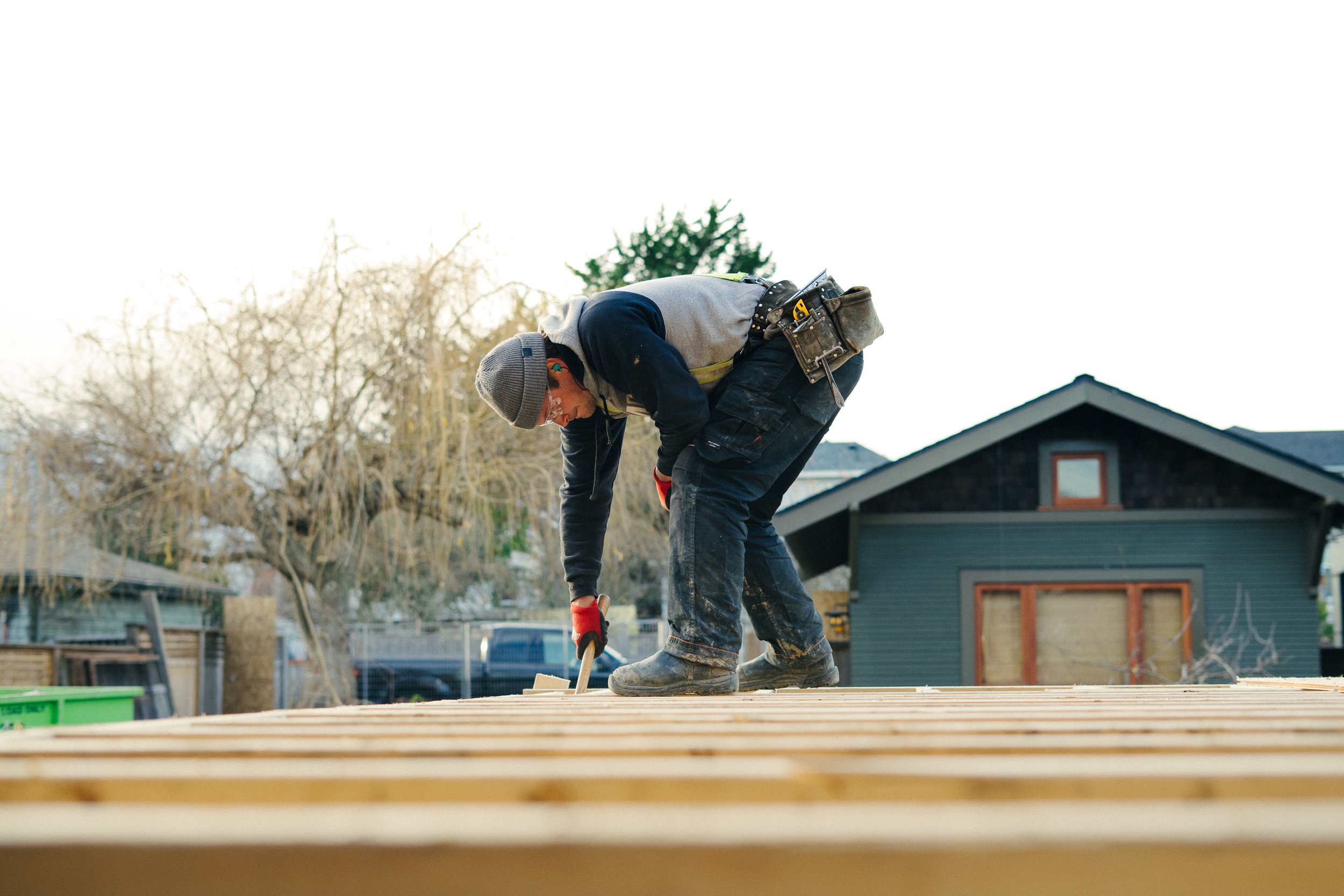 A construction worker is bent over, hammering nails into a wooden roof frame outdoors during daytime.