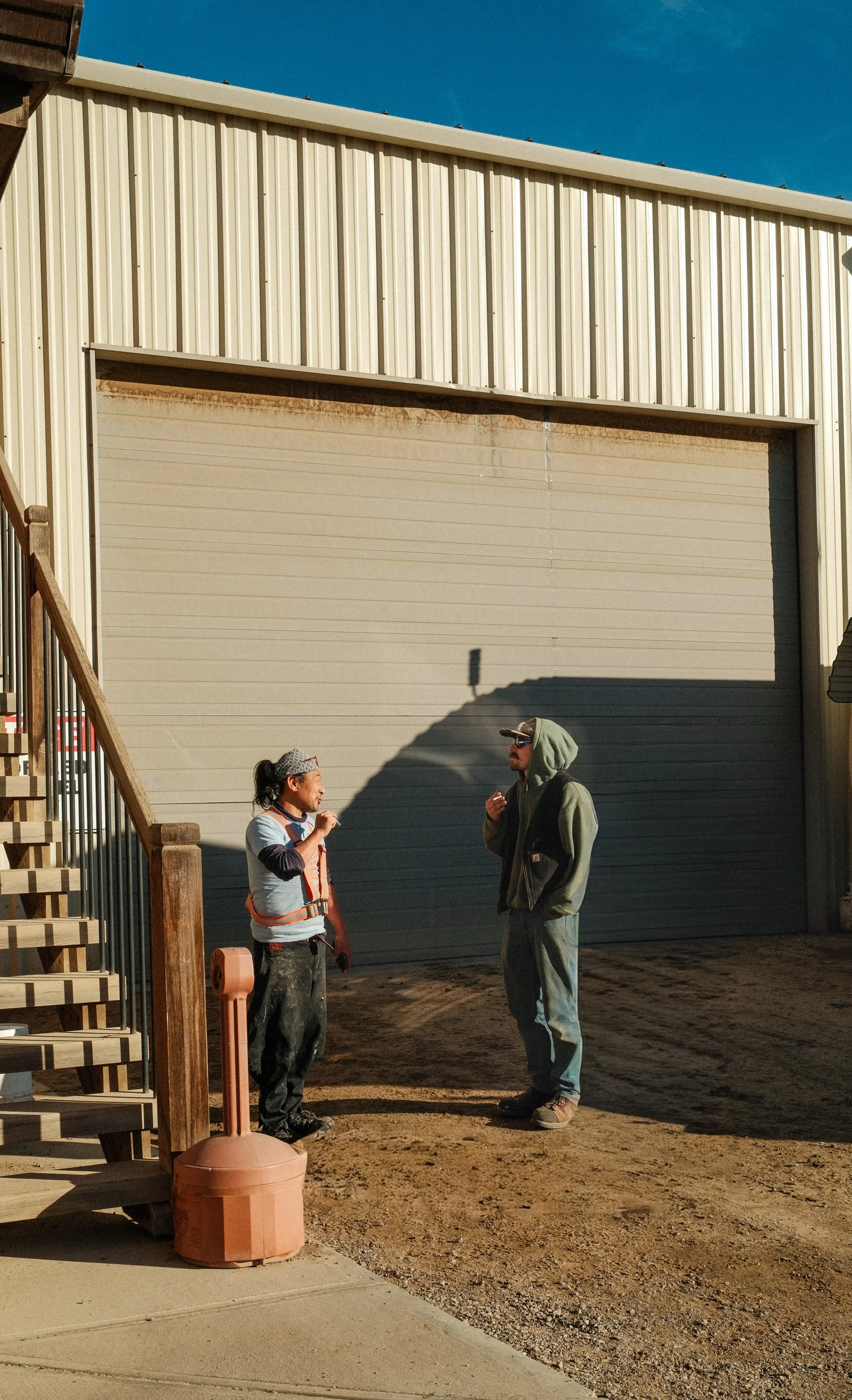 Two men are standing and talking in front of a large garage door outside a building on a dirt surface. One man is wearing a hoodie, sunglasses, and jeans. The other man is wearing work clothes, a headband, and appears to be a worker. Shadows cast on 