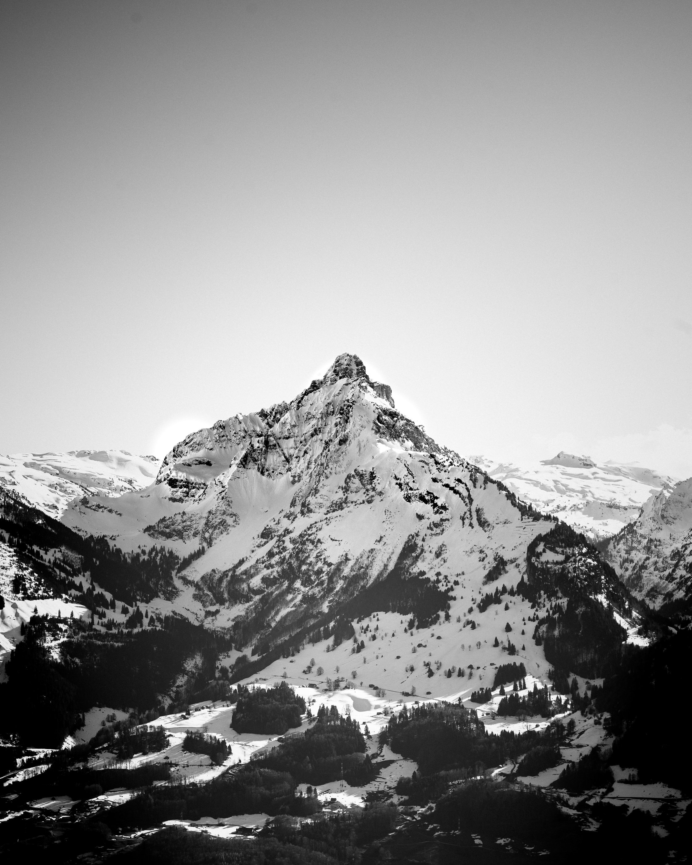 Black and white photograph of a snow-covered mountain with a rugged peak, surrounded by smaller snow-covered hills and forests.