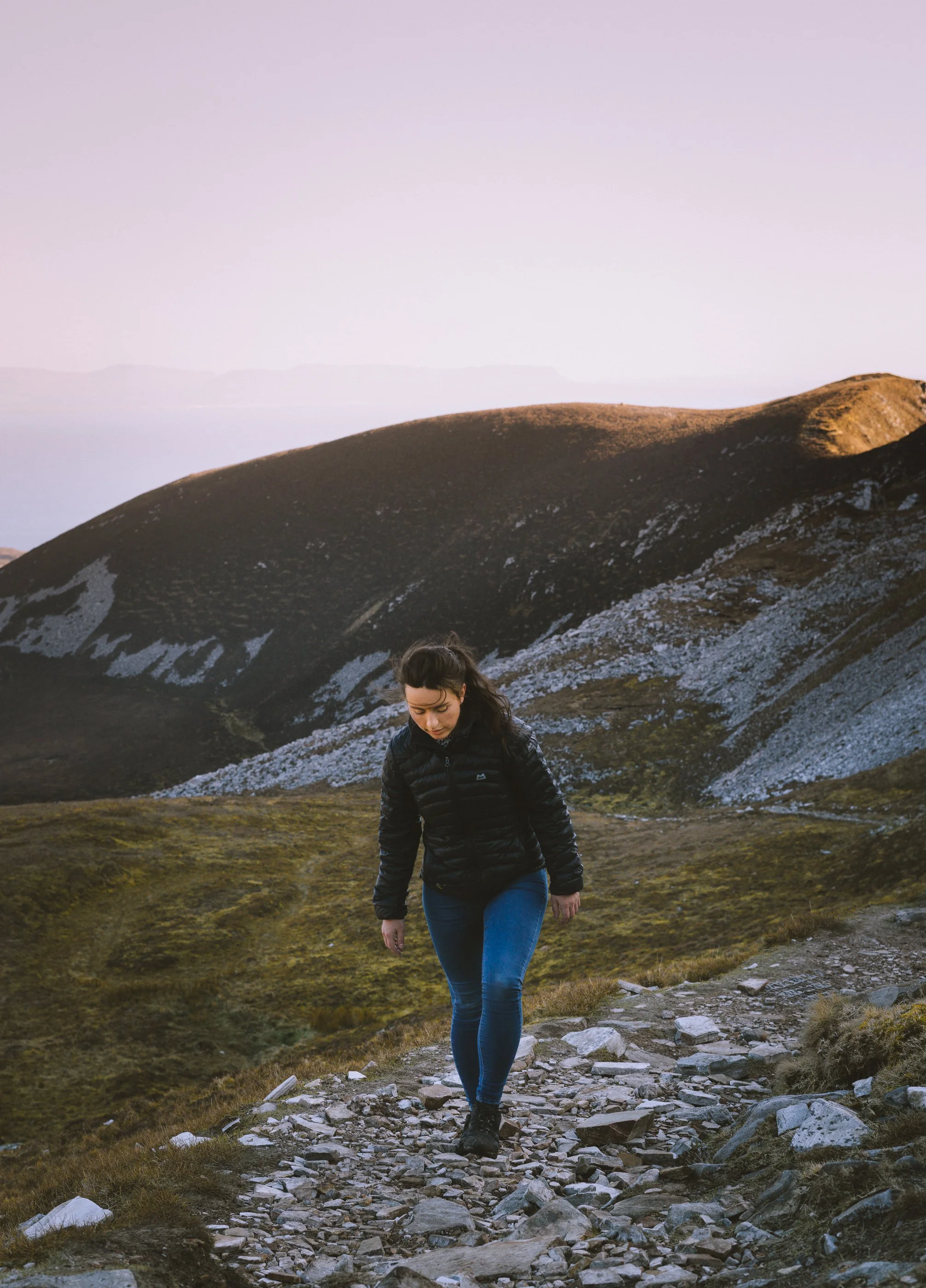 A woman in outdoor hiking gear walking on a rocky trail with mountains and hills in the background during dusk or dawn.