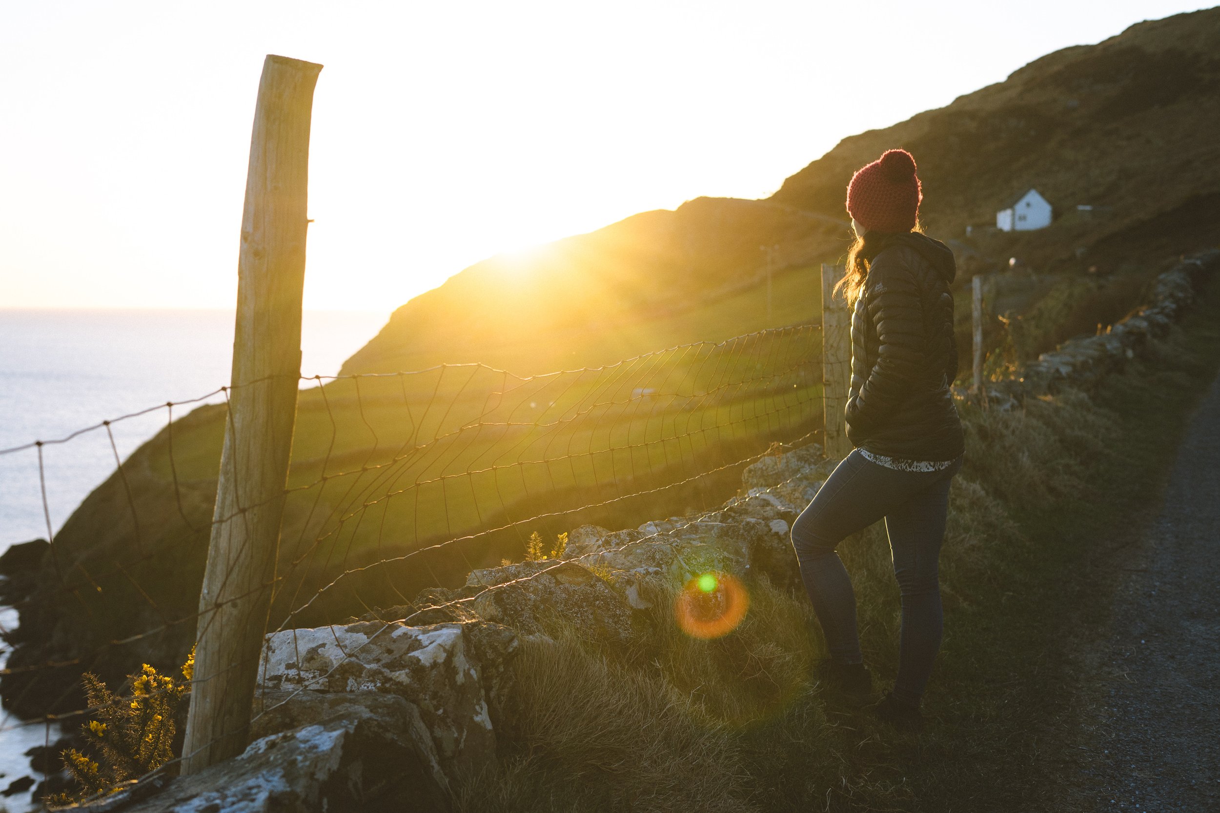 Woman standing on a coastal path with grass and rocks, wearing a red knit hat and black jacket, looking toward the sunset over the ocean and hilly landscape.