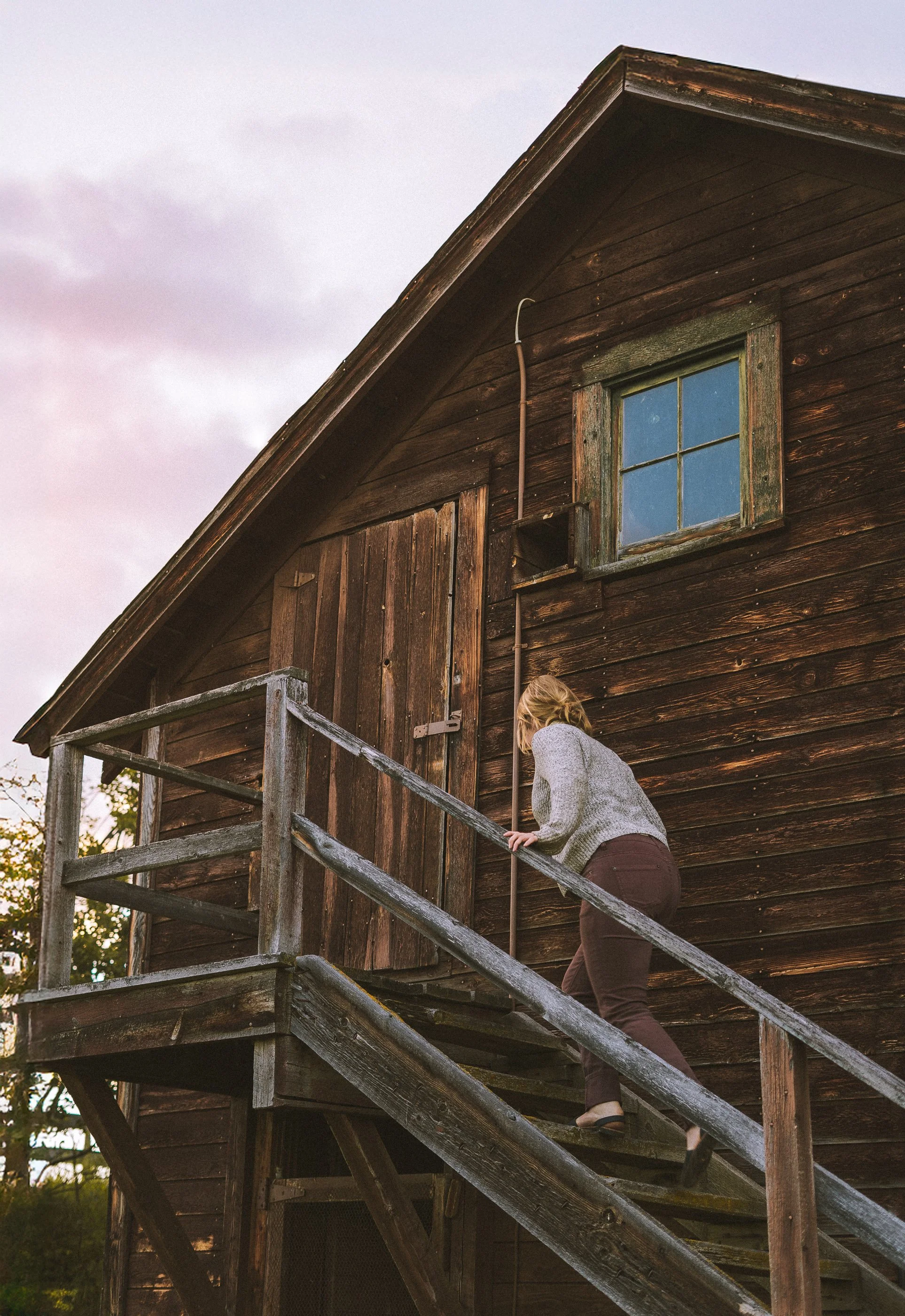 A woman with blonde hair wearing a gray sweater and brown pants climbing wooden stairs towards an old rustic wooden barn with a small window and a closed door.