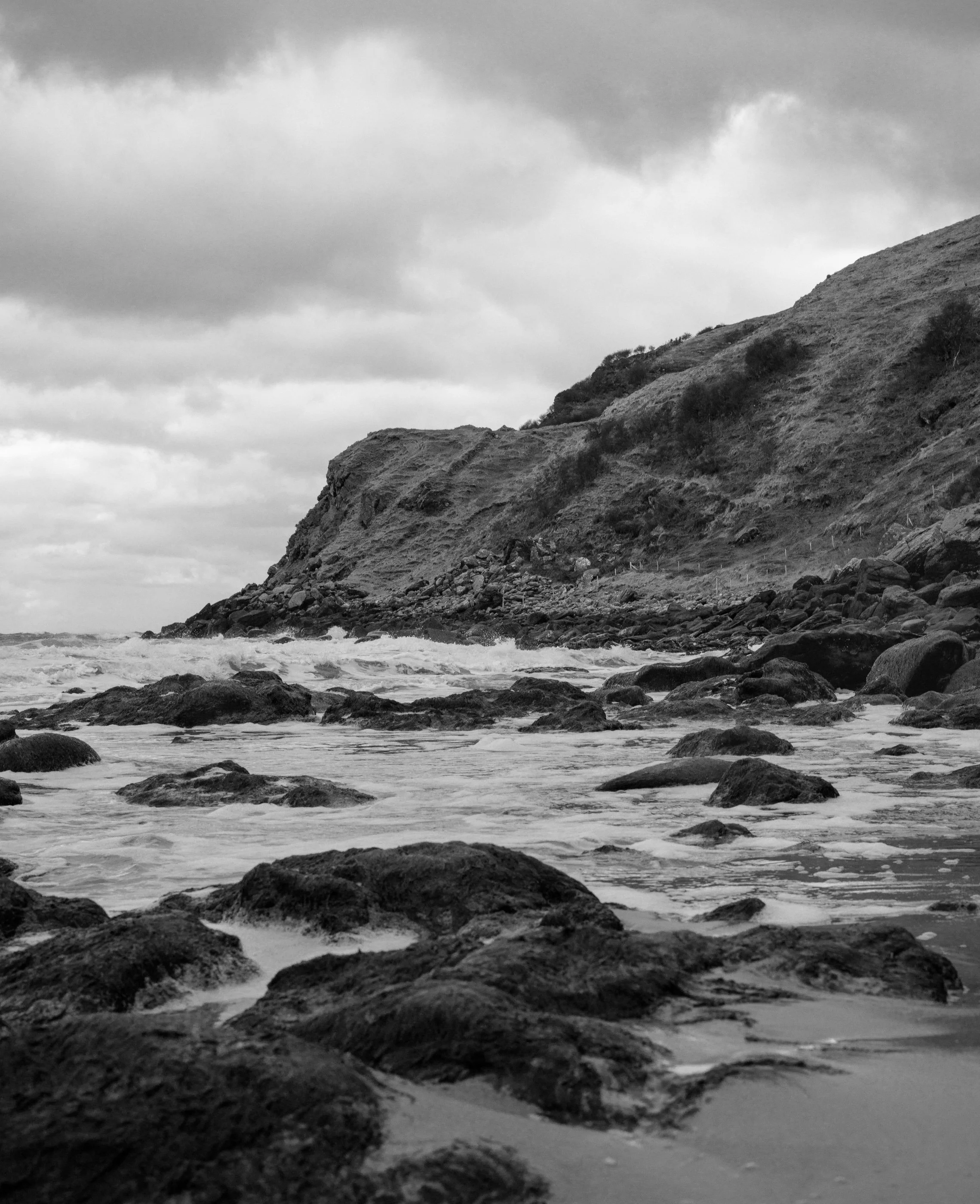 Black and white photo of a rocky beach with waves crashing against rocks and a hillside in the background, cloudy sky above.