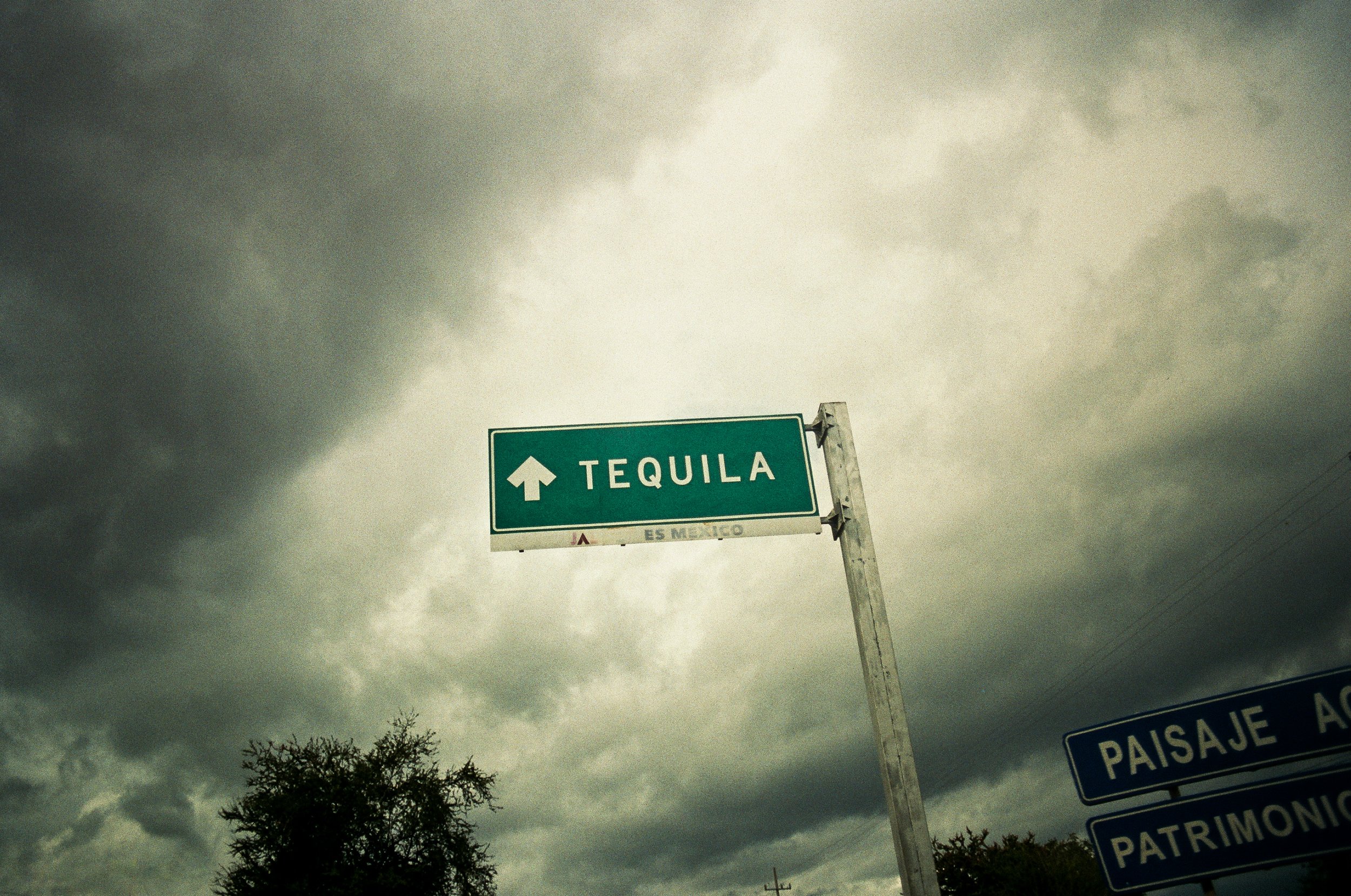 Green road sign with white text and arrow pointing upward that says 'TEQUILA' against a cloudy sky.