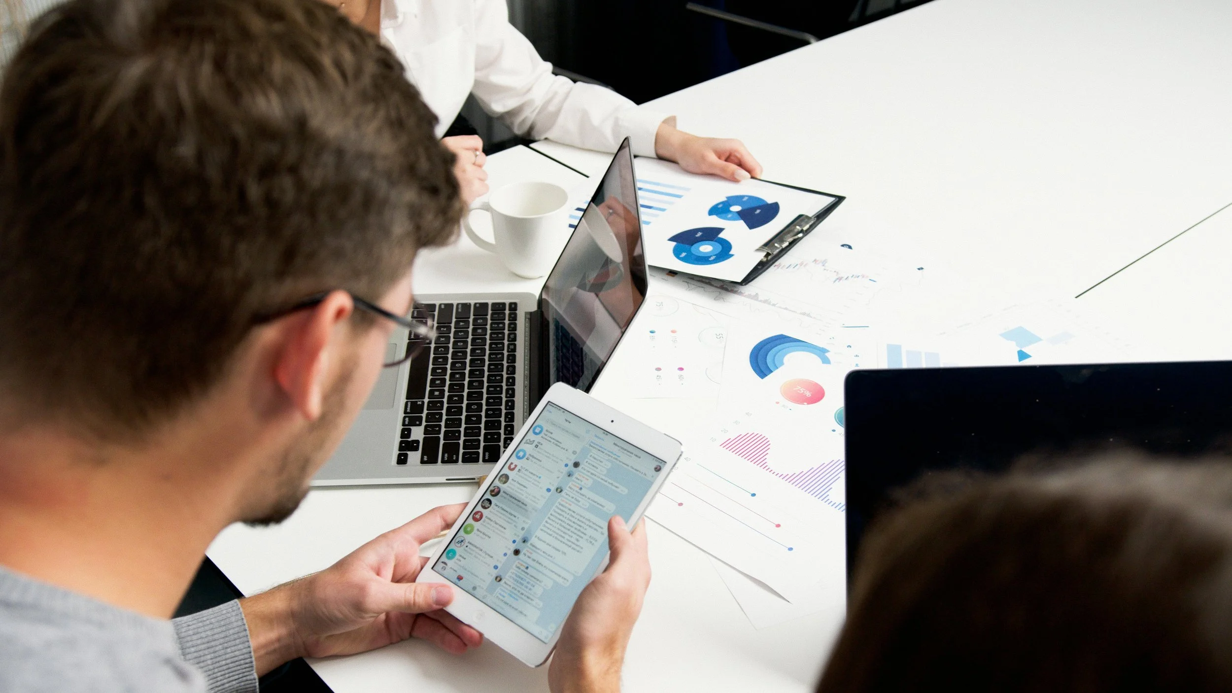 A man sits at a conference table between 2 female coworkers. He holds a tablet and reviews messages. Several printed graphs lay on the table and 2 laptops are nearby.