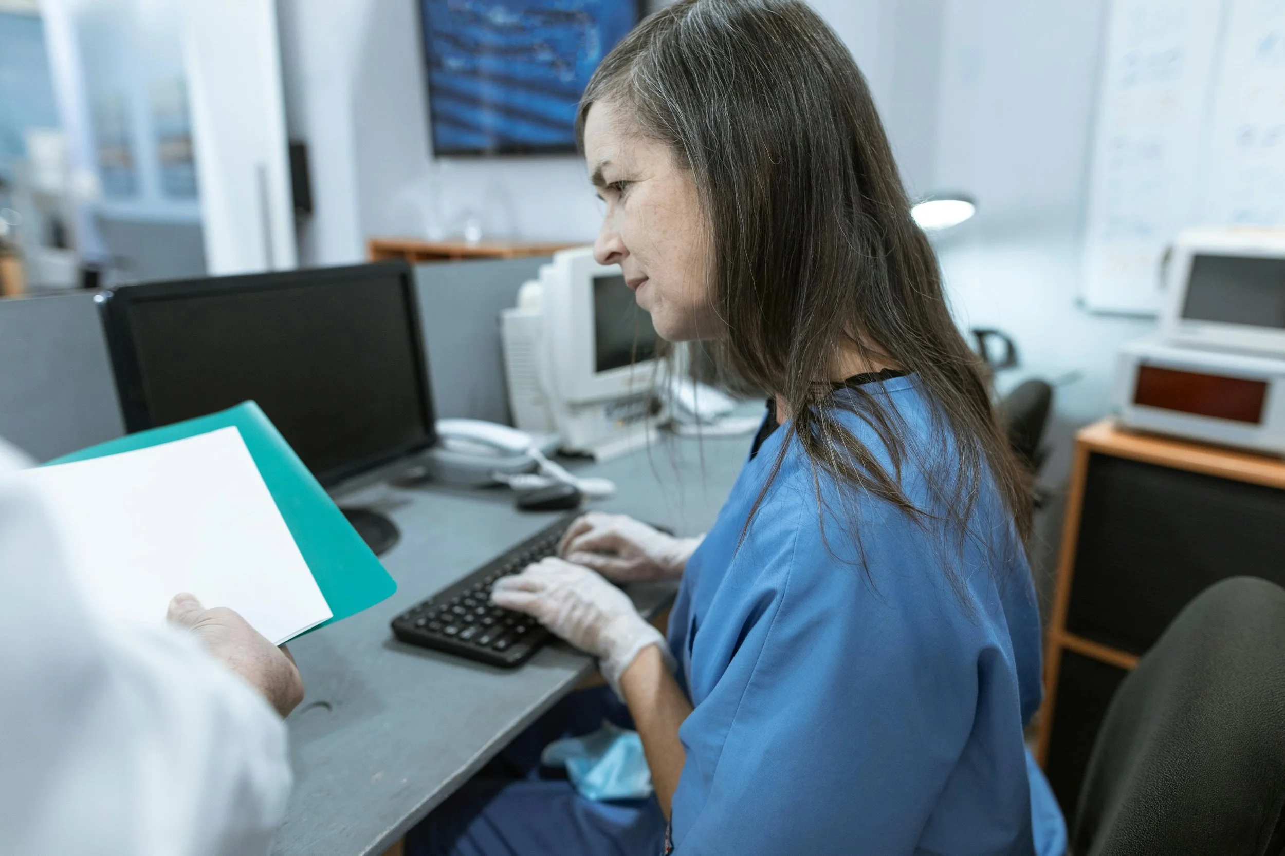 A female medical provider, wearing blue scrubs stands at a desk. Her gloved hands hover over a computer keyboard as she reviews paper documents held by an out of frame individual.
