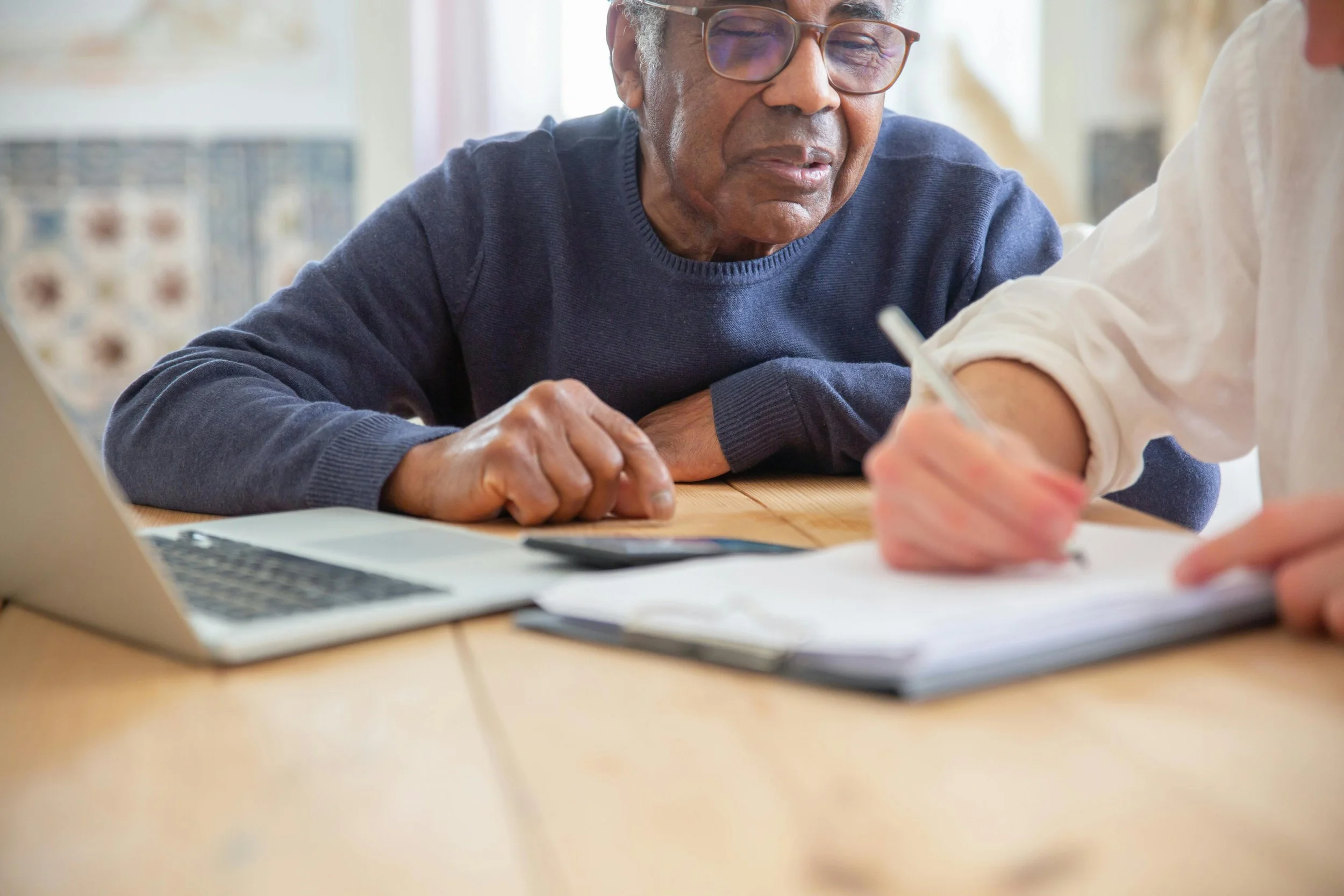 An older man in a blue sweater sits at a table in his home. He is watching a medical provider take notes on his health status.